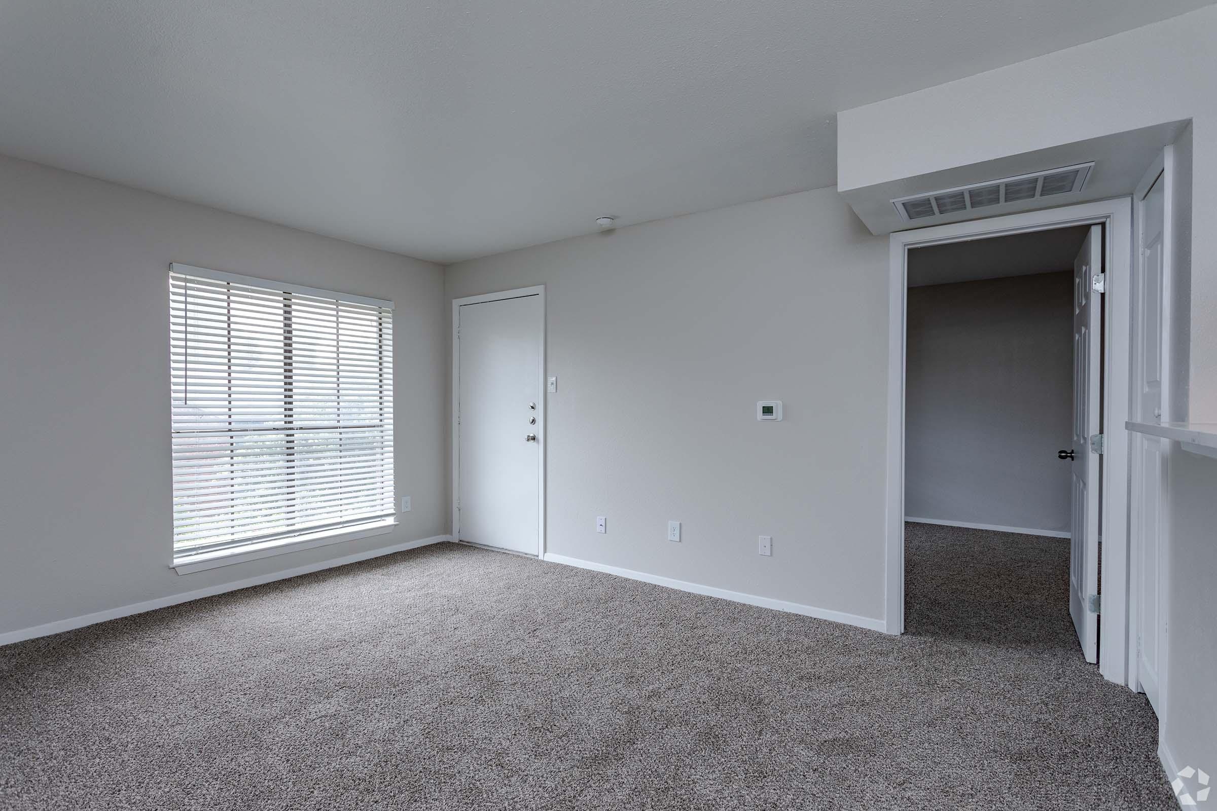 A spacious, empty room with light gray walls and plush carpeting. A window with white blinds allows natural light to enter. There is a door leading outside, and another door visible in the background, suggesting a layout that connects to other areas. The overall ambiance is clean and neutral, ideal for personalization.