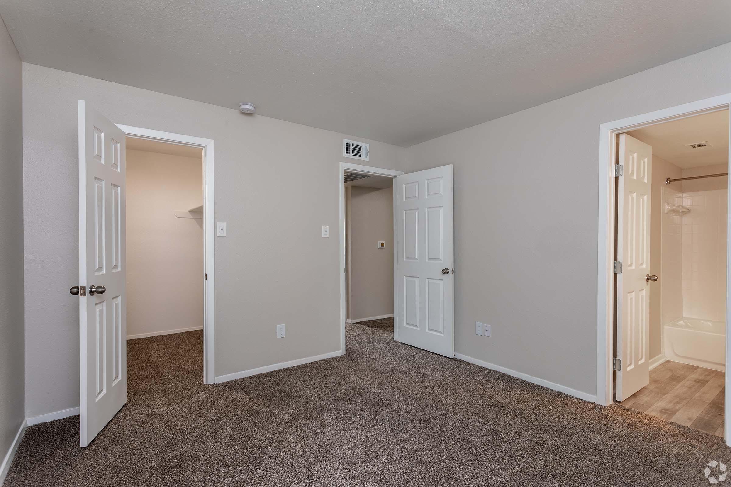 A well-lit, empty room with light beige walls and carpeted flooring. There are three white doors: one opened to a closet, another leading to a bathroom with a shower, and a third door closed. The room offers a spacious, clean look, suitable for various uses.