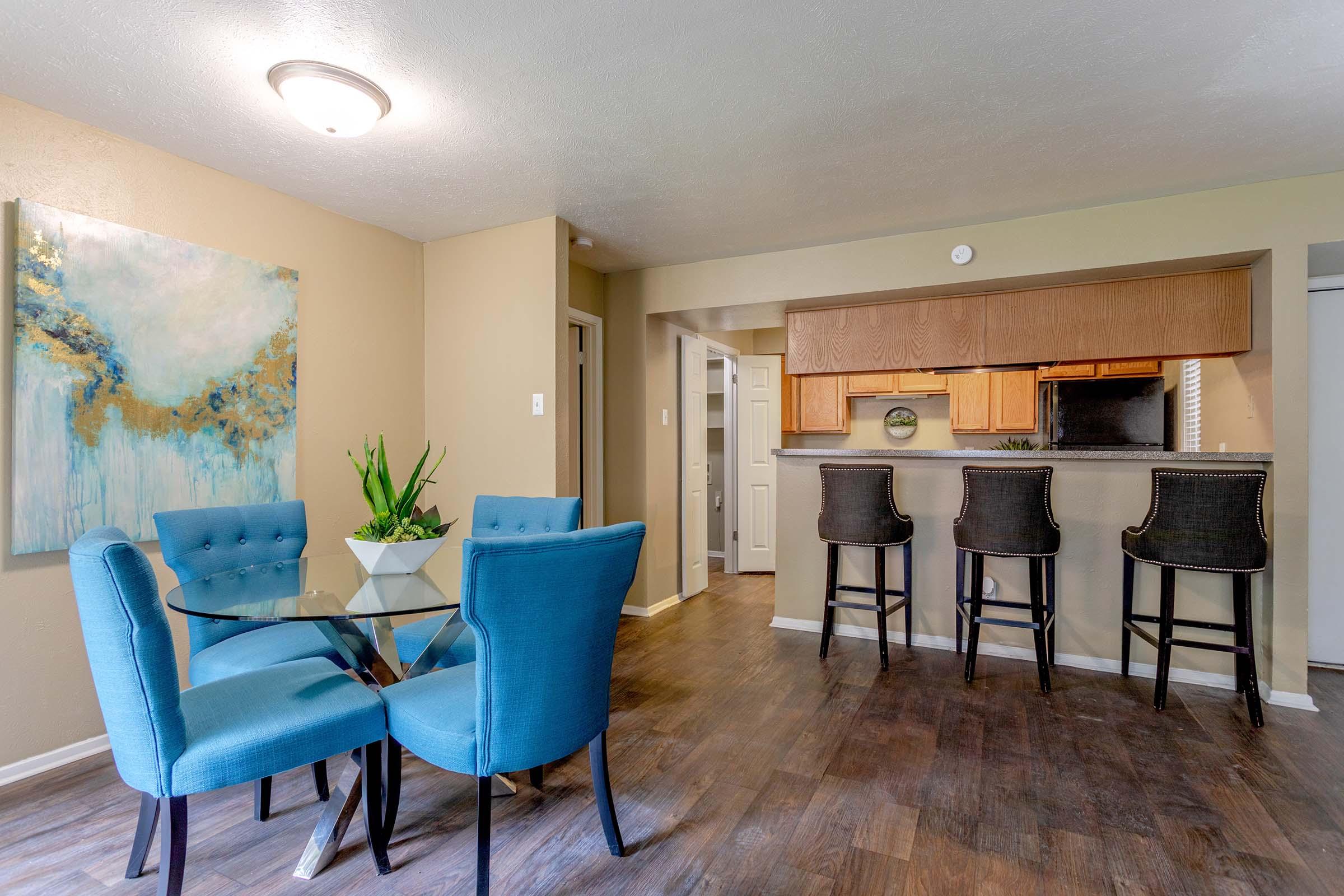 A modern dining area with a glass table and four turquoise chairs. In the background, there is a kitchen bar with three high stools. The walls are painted in a warm, neutral tone, and there's a large piece of abstract wall art featuring blue and gold colors. Natural light fills the space.