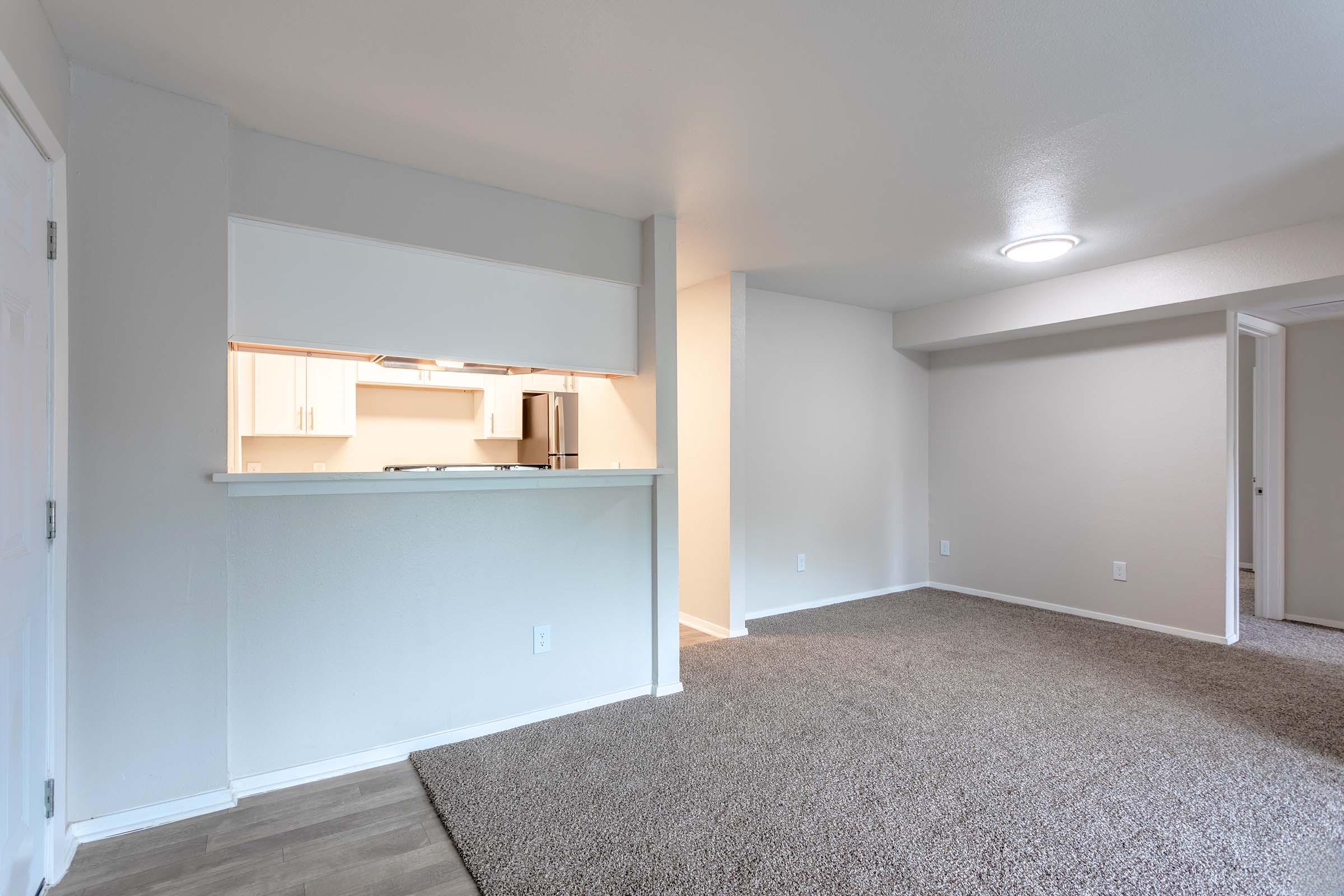 A spacious, modern living area featuring light beige carpet and neutral-colored walls. An open kitchen with a breakfast bar is visible in the background, showcasing white cabinetry and stainless steel appliances. Soft, natural light illuminates the room, enhancing its inviting atmosphere.