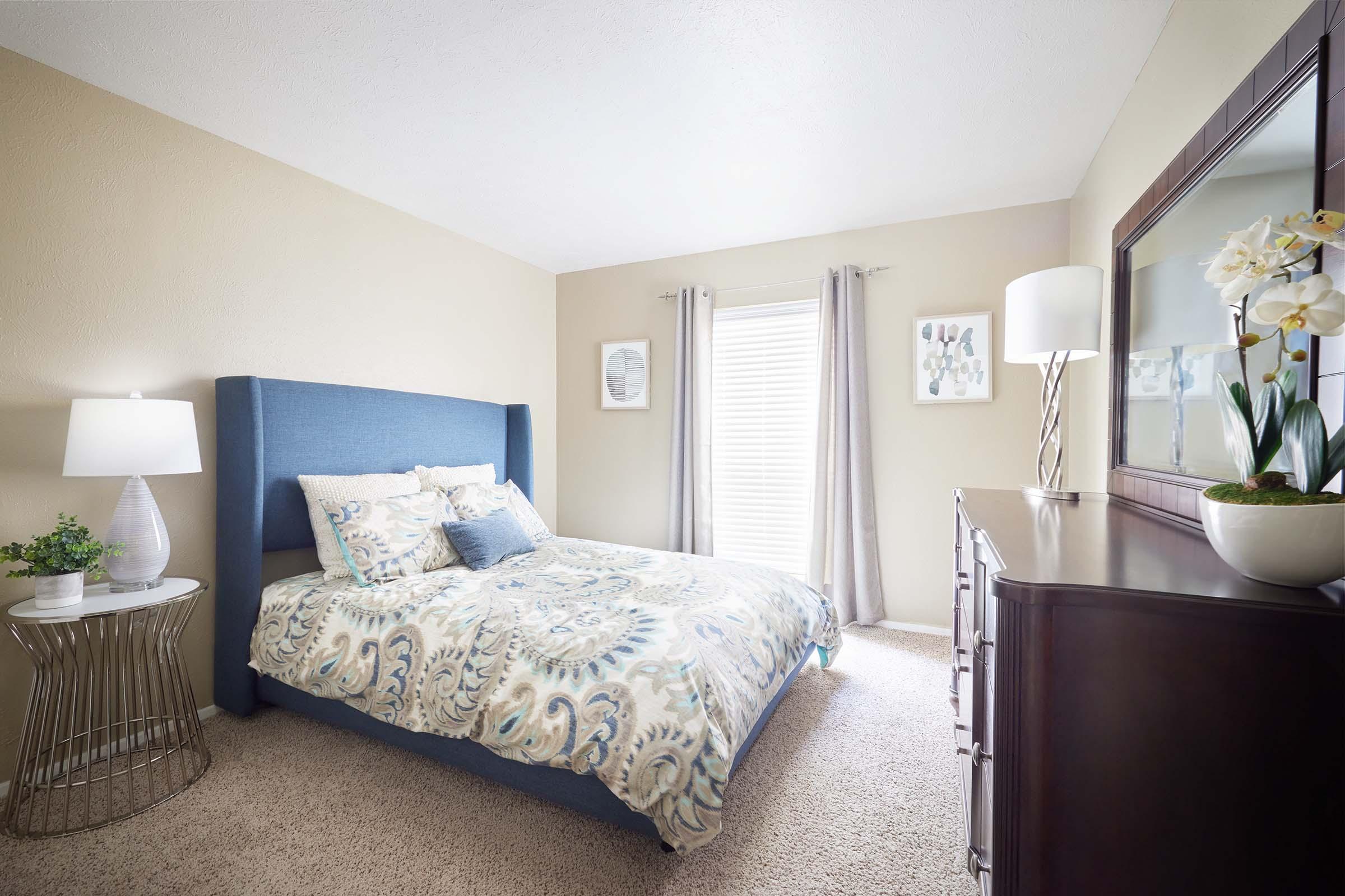 Cozy bedroom featuring a blue upholstered bed with a decorative comforter, accented by pillows. A bedside table with a lamp and a potted plant is on one side, while a dark wooden dresser and a mirror complement the space. Natural light filters through sheer curtains, creating a warm atmosphere.