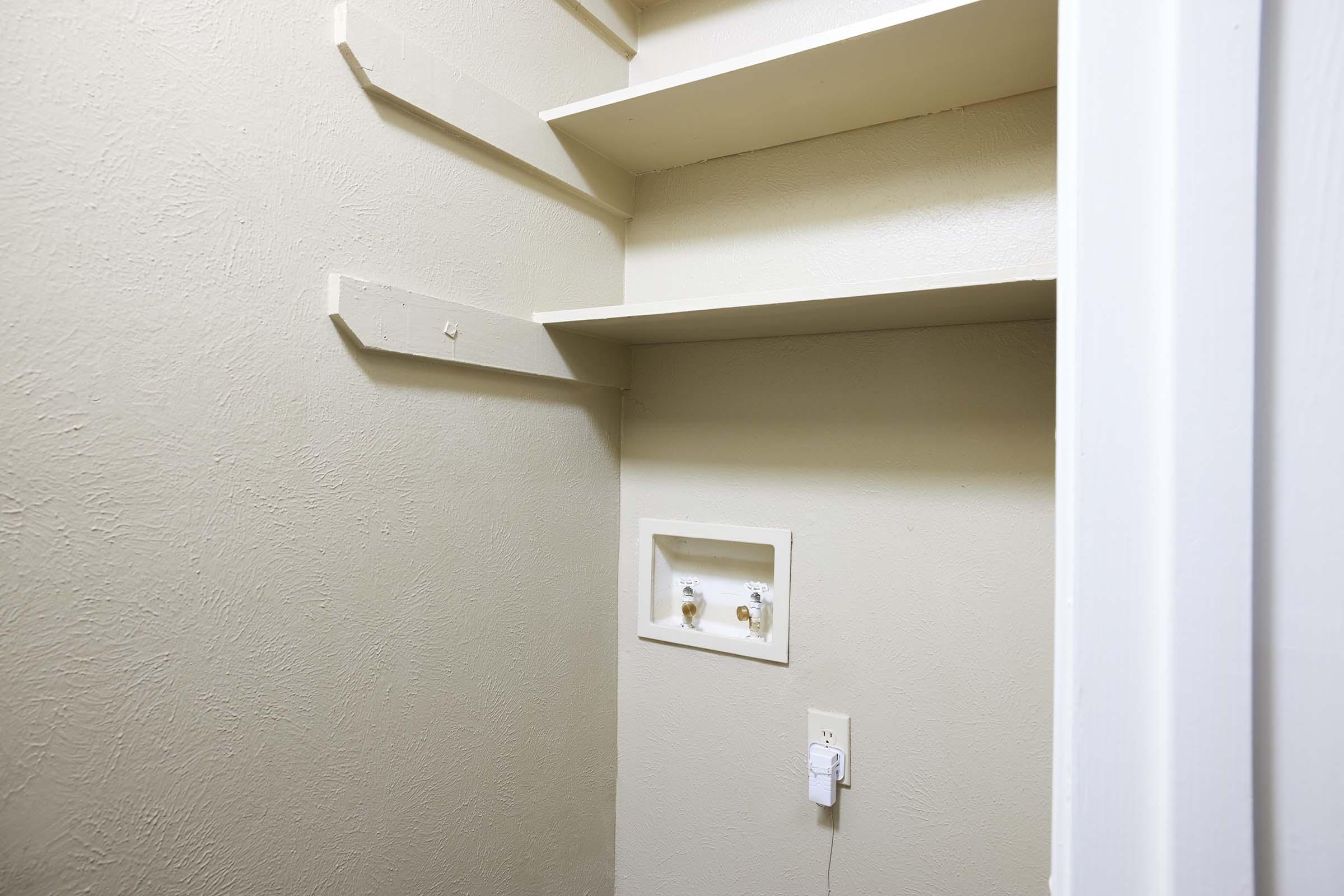 A sparsely furnished room with light beige walls and a couple of shelves attached to one side. There is a small panel with two pipes and a power outlet on the wall. The space appears empty and unadorned, suggesting it may be a utility or storage area.