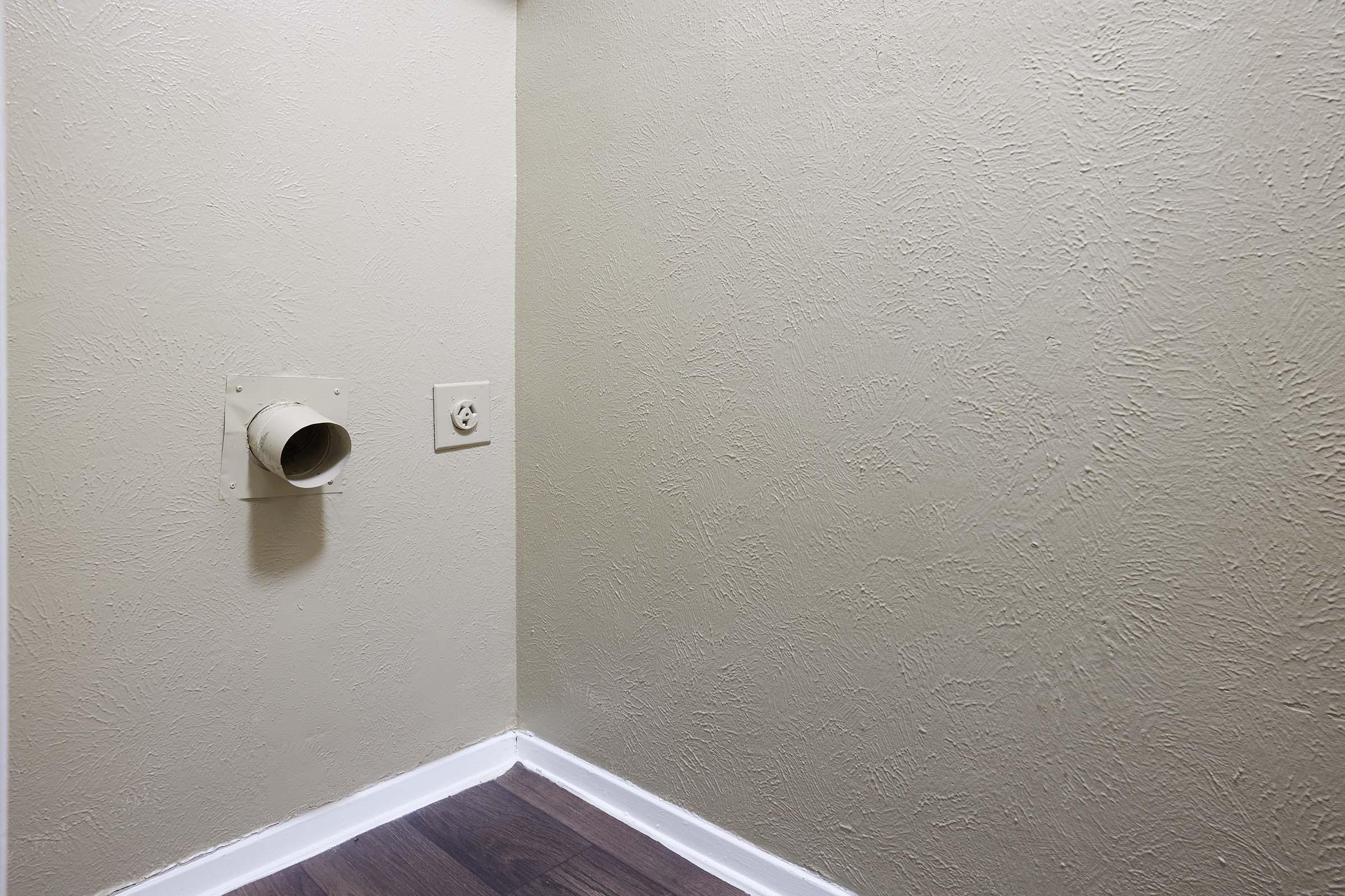 A clean corner of a room with a beige textured wall. On the left, there is a round vent leading outside, and on the right, an electrical outlet. The floor has a dark wooden finish, and the baseboard is white, creating a simple, uncluttered look.
