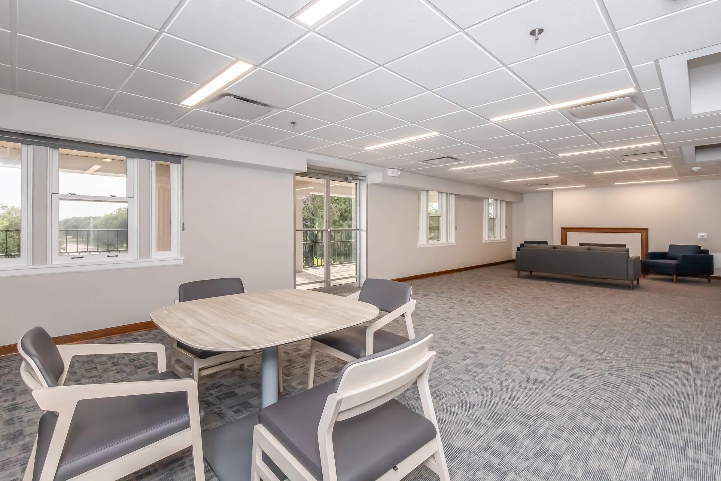 A spacious, modern interior room with a light-colored carpet. There is a round wooden table surrounded by four chairs, and a dark sofa in the background. Large windows provide natural light, and double doors lead outside. The overall decor is minimalistic and contemporary.