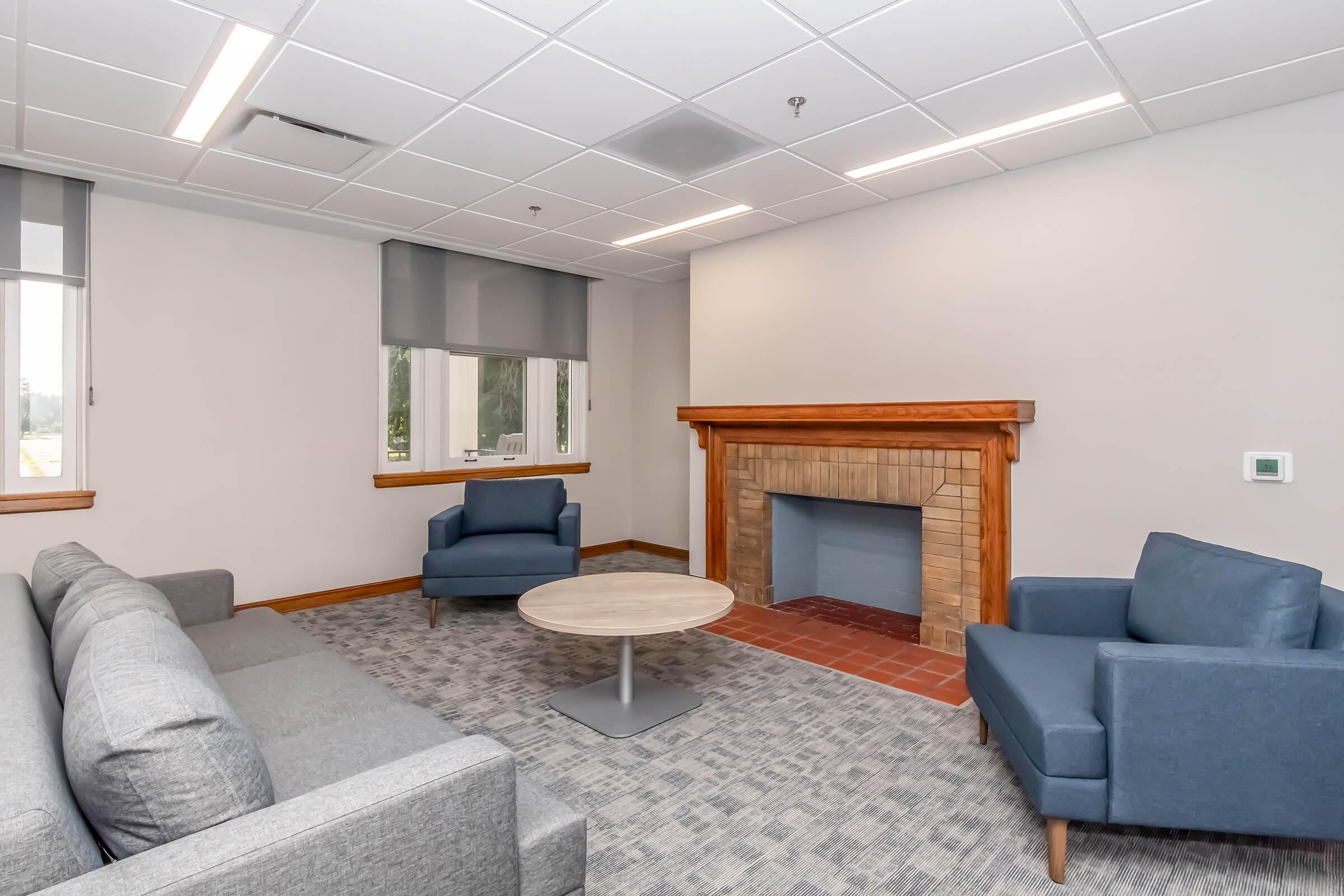 A modern lounge area features a gray sofa and two blue chairs arranged around a circular table. A fireplace with a brick surround is situated against the wall, and natural light streams in through two windows with roller shades. The floor is covered with a light gray carpet.