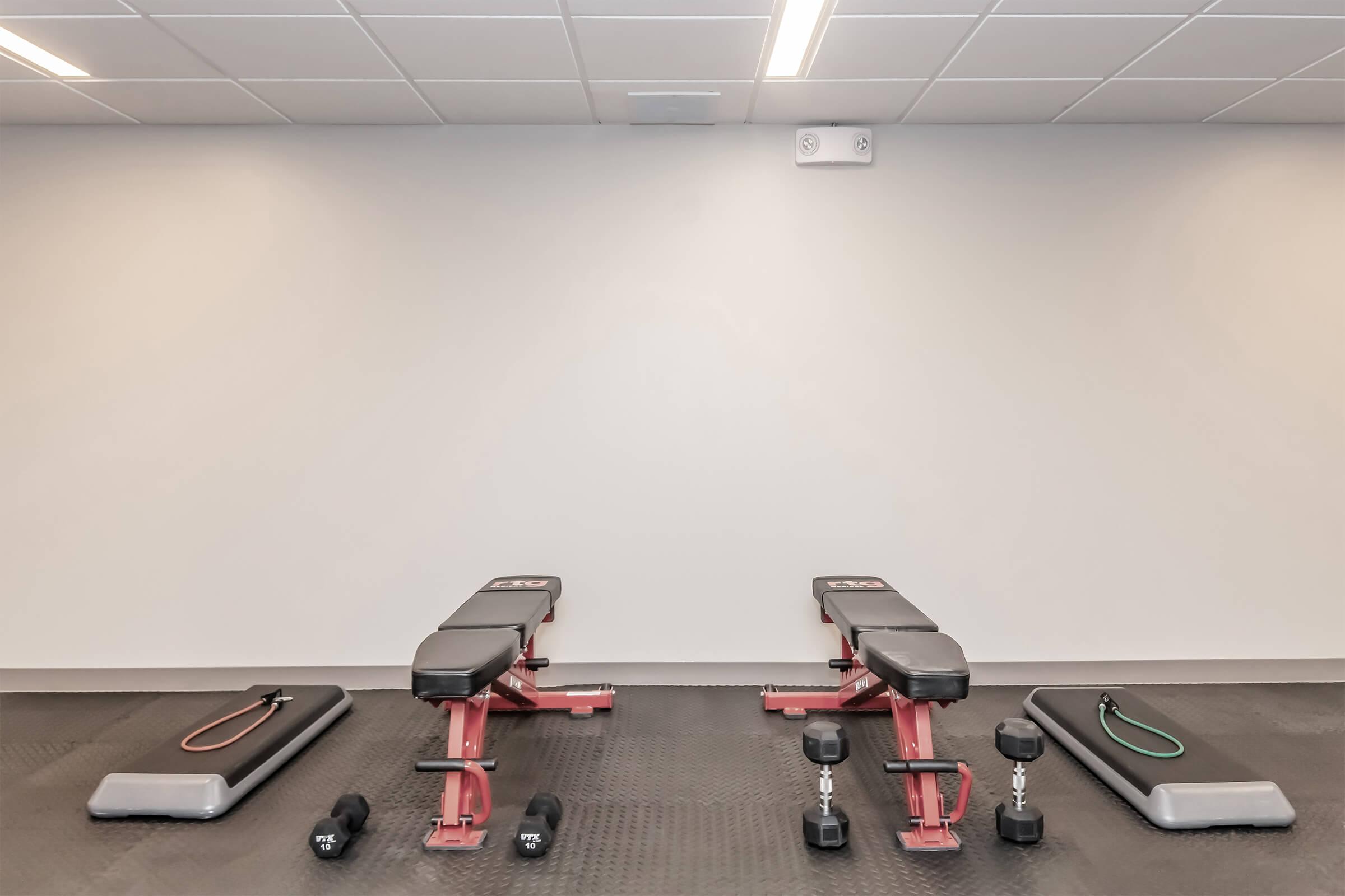 A clean gym space featuring two workout benches, two sets of dumbbells, and resistance bands laid out on a black rubber floor. The background is a plain, light-colored wall with overhead lighting.