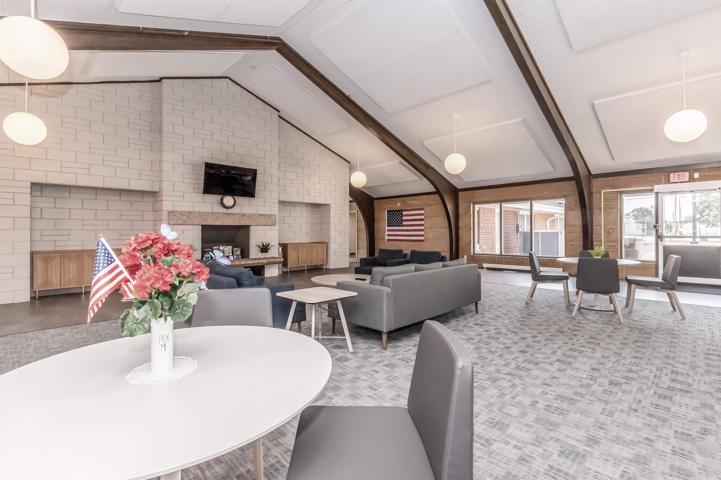 A spacious modern lounge area featuring a large fireplace, comfortable seating with gray sofas, a round table with chairs, and decorative elements including a vase of flowers and an American flag. Large windows allow natural light to fill the room, enhancing the inviting atmosphere.