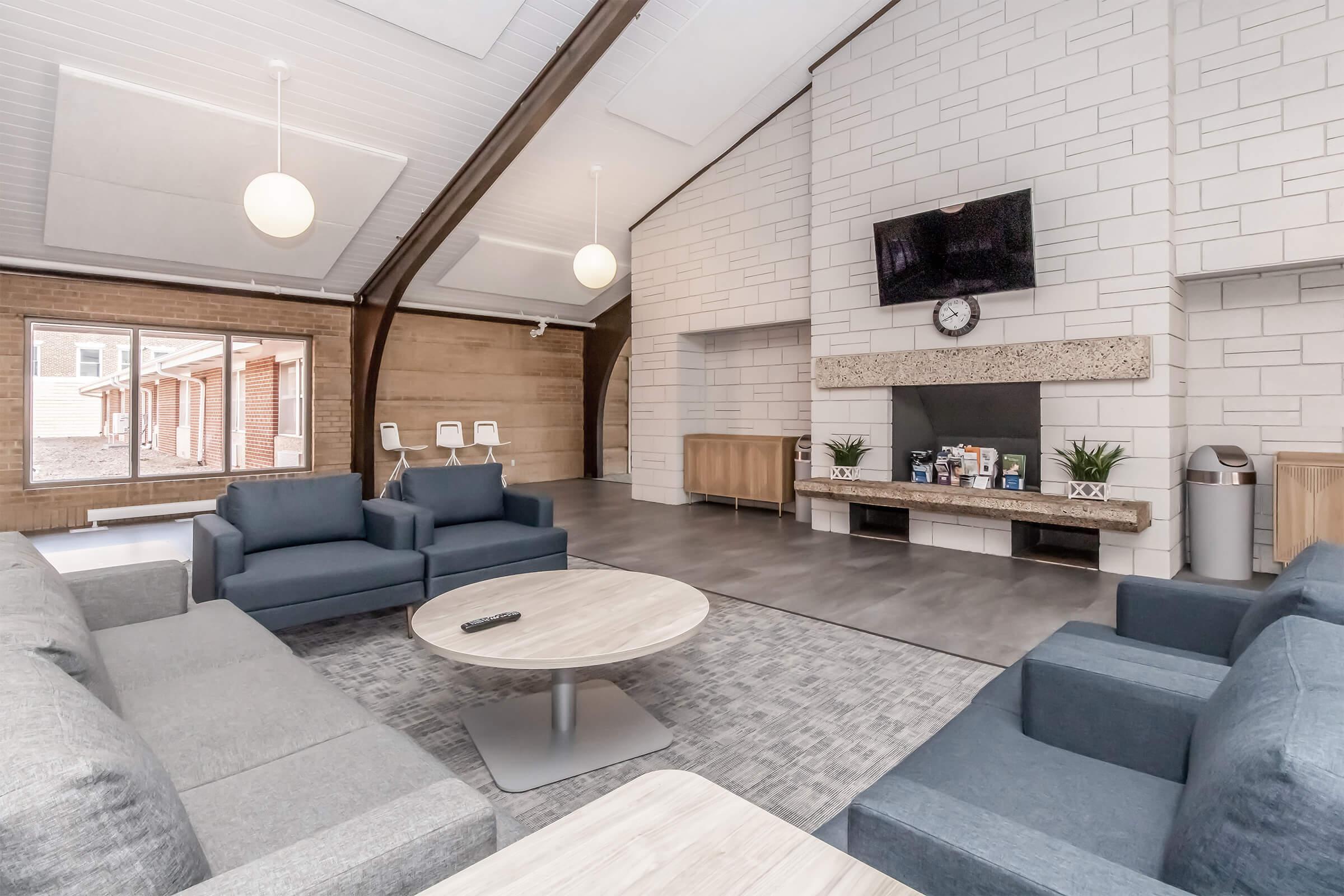 A spacious modern lounge area with grey sofas and a round table. A large flat-screen TV is mounted on a stone feature wall, above a shelf with decorative items. Large windows allow natural light to fill the room, and a trash bin is visible in the corner. The design features high ceilings and wooden elements.