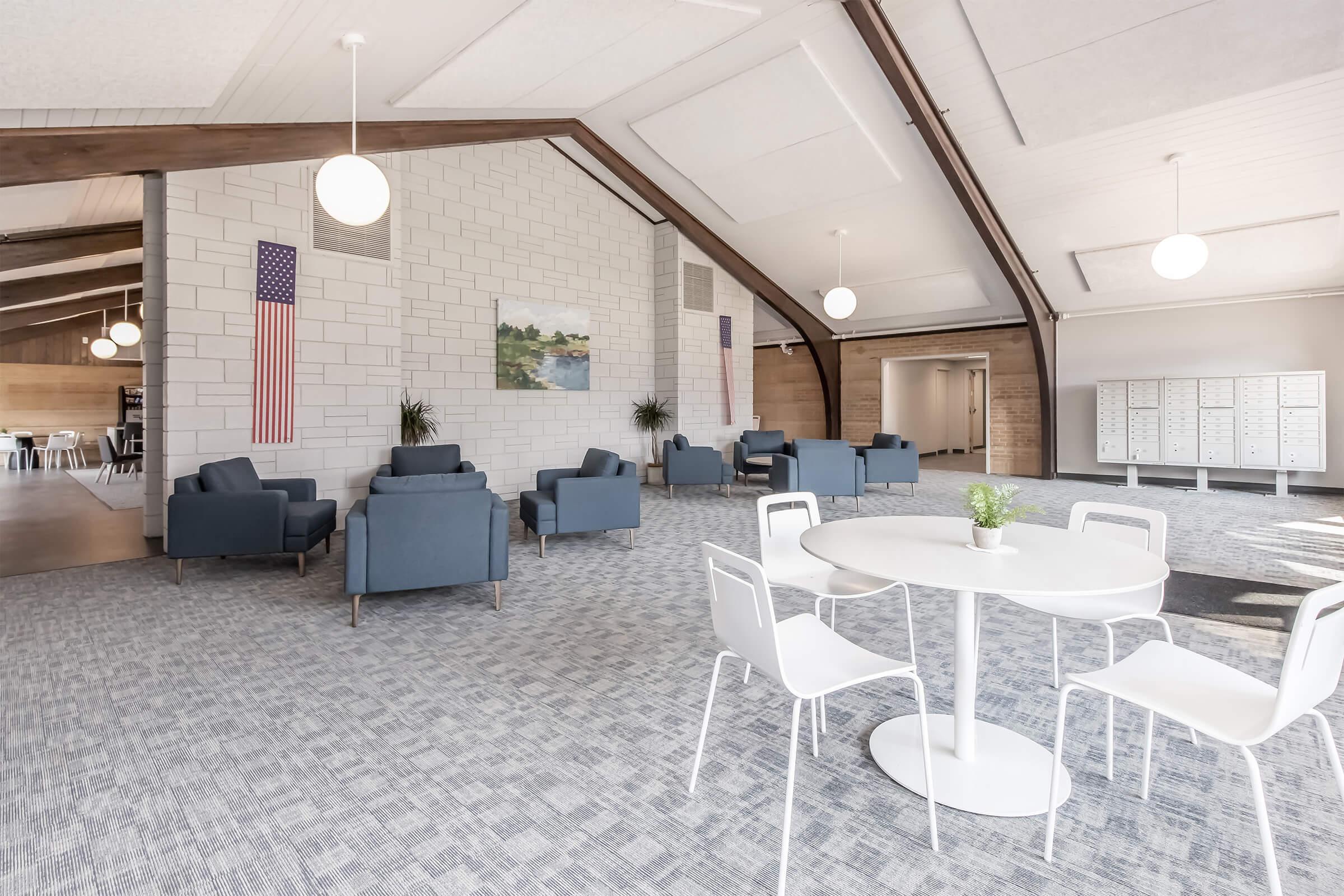 Spacious indoor area featuring modern furnishings, including blue chairs and a round table. Walls are light-colored, with wooden beams and large windows. An American flag hangs on the wall, and there’s a painting of a landscape. Lockers are visible in the background, adding to the functionality of the space.