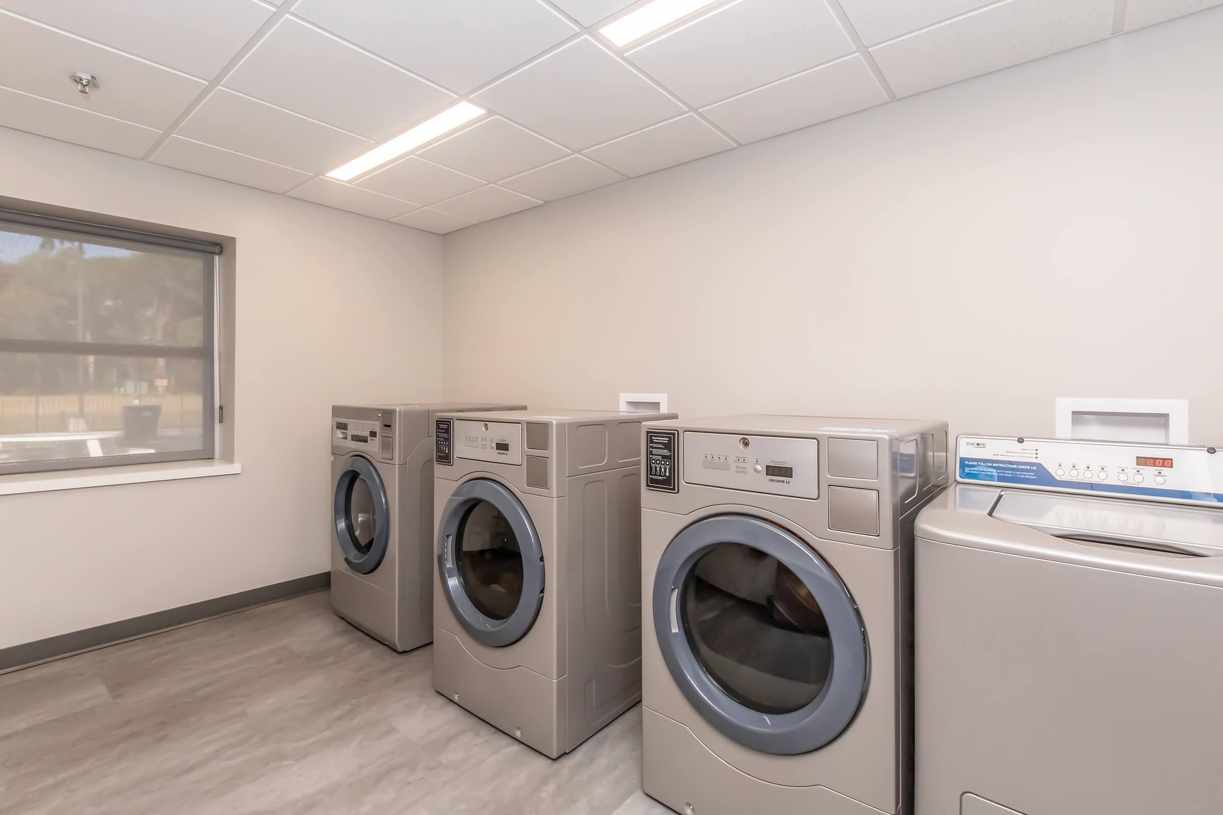 A clean laundry room featuring four large washing machines and a dryer. The machines are metallic gray and are lined up against a light-colored wall. A window allows natural light to enter, enhancing the bright and modern atmosphere of the space. The floor is finished with a light-colored material.