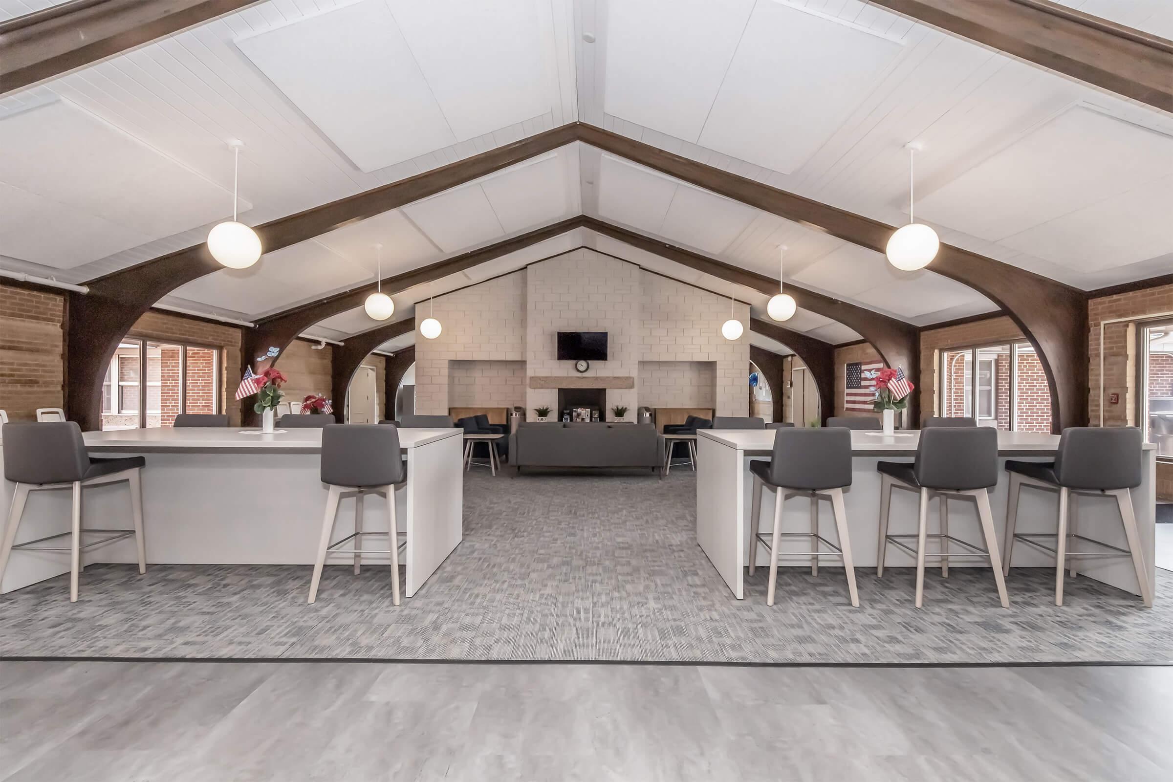 A spacious interior of a modern common area featuring a high ceiling with wooden beams, light fixtures, and a cozy seating arrangement. There are bar-style tables with chairs on one side and a fireplace with a television above it at the far end. Large windows provide natural light.