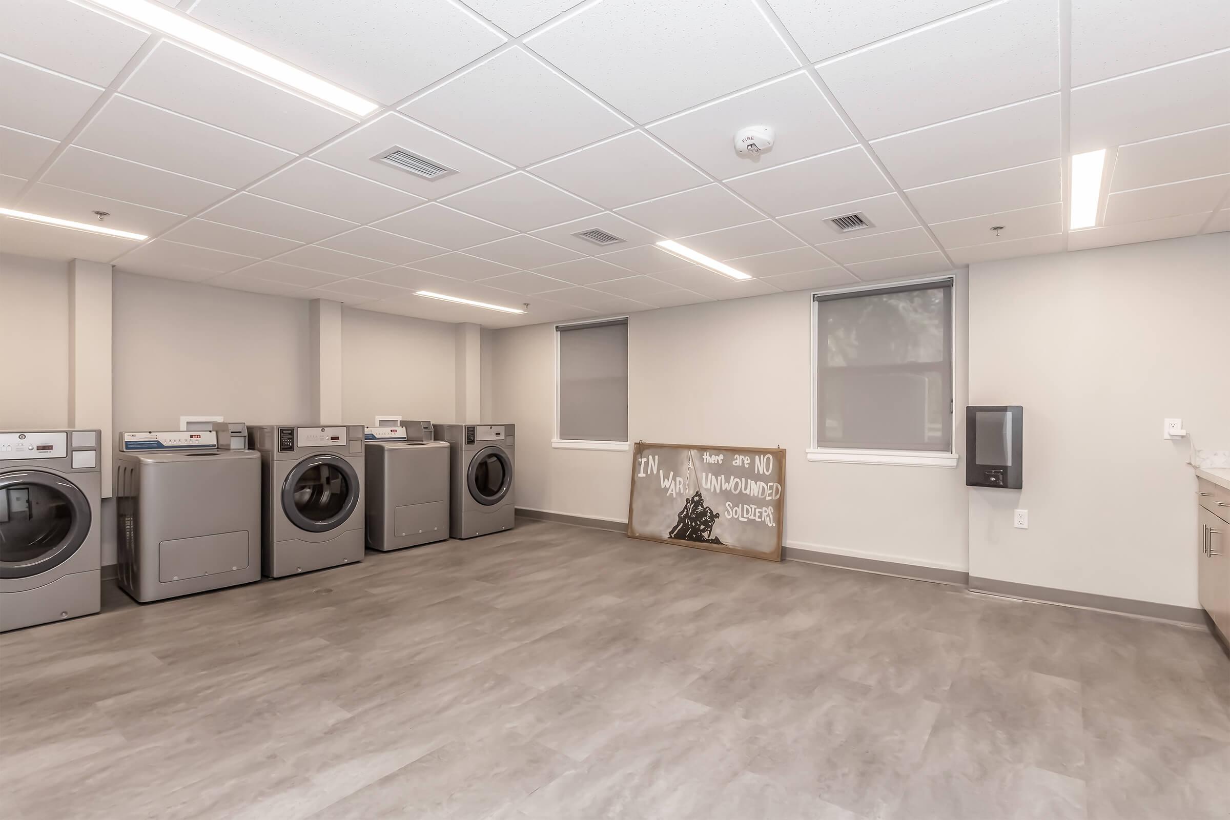 A spacious laundry room featuring several washing machines, a large window with shades, and a sign on the floor that reads "I'M A MAN. UNWOUNDED SOLDIER." The room has modern lighting and a clean, minimalist design with gray flooring.