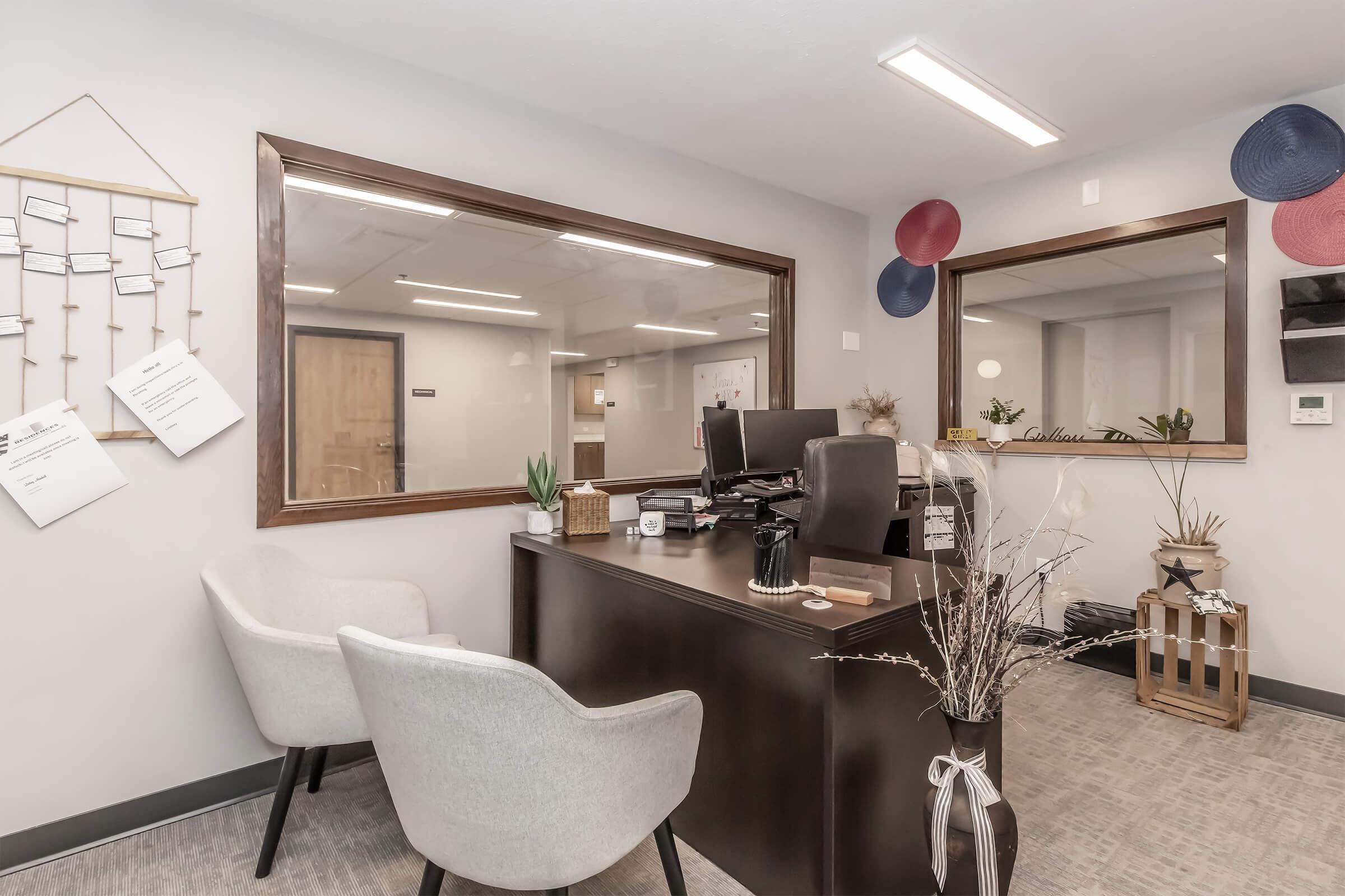 An office workspace featuring a reception area with a dark wooden desk, two comfortable white chairs, and modern decor. There are glass windows allowing visibility into another office space, adorned with plants and decorative items. Papers are displayed on the wall, adding to the organized atmosphere.