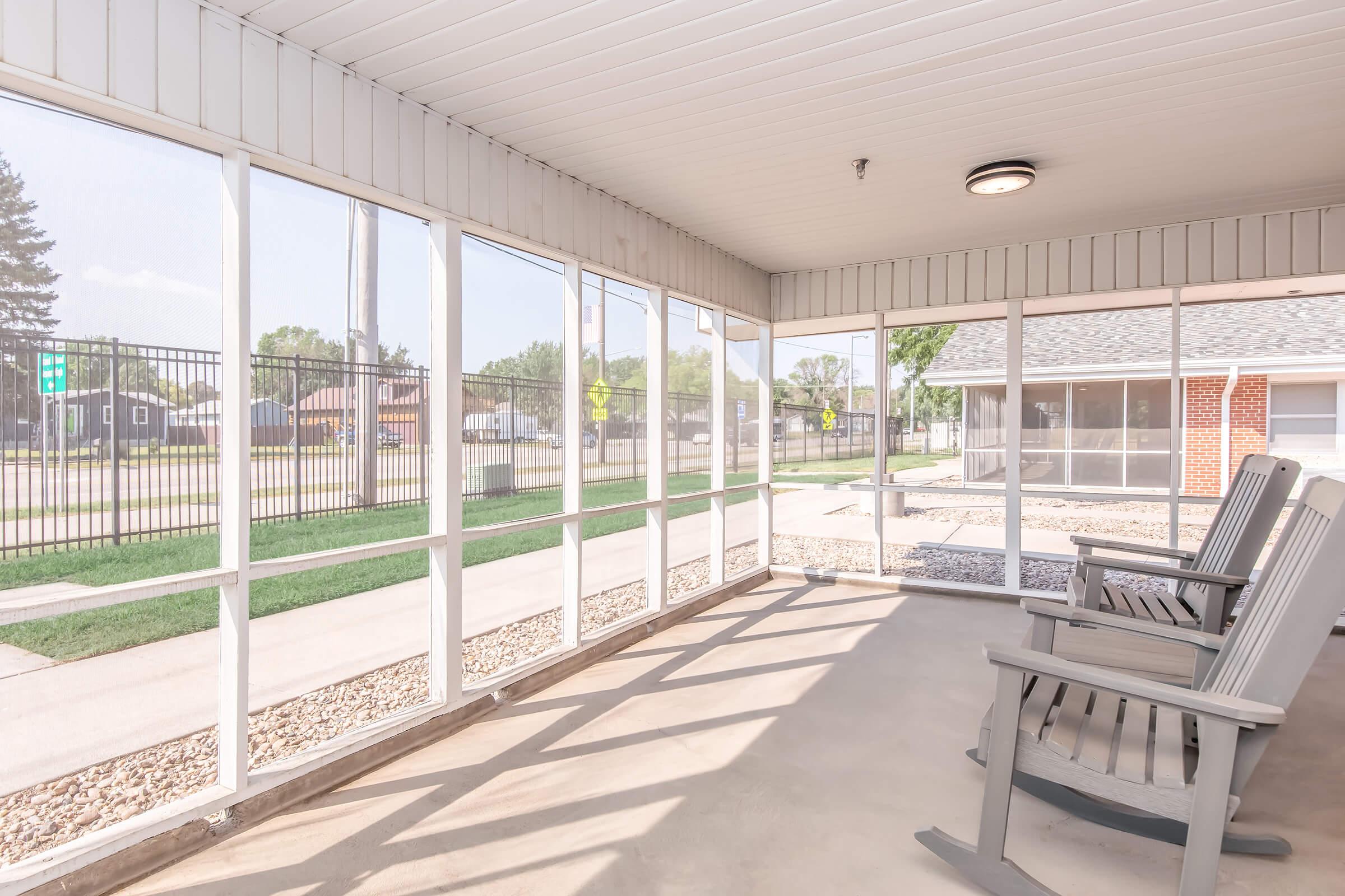 A bright and airy screened porch featuring several rocking chairs, with sunlight streaming in through the screens. The flooring is concrete, and there are visible outdoor elements, including grass and a fence in the background. The area is designed for relaxation and enjoying the outdoors while being sheltered.
