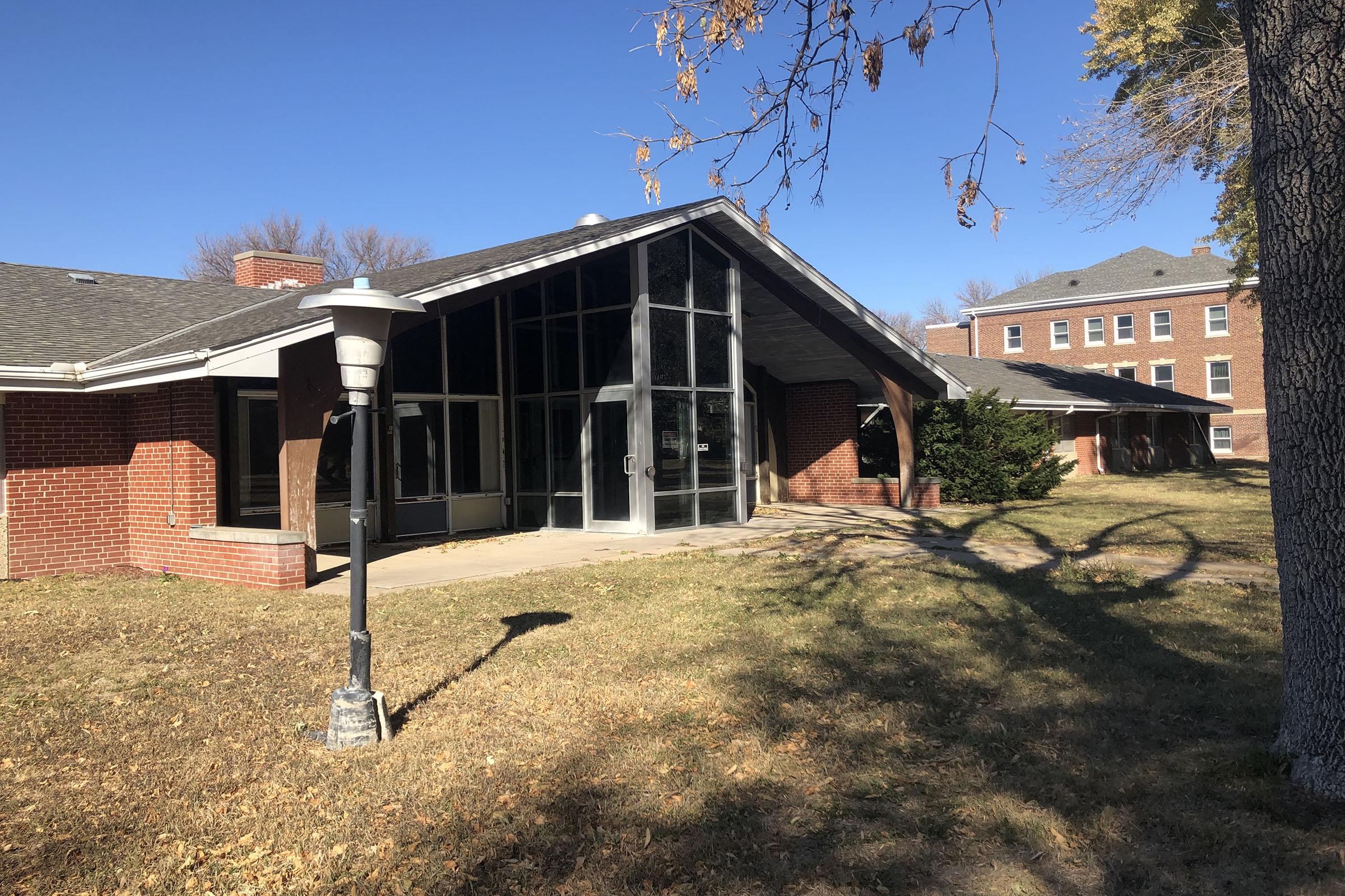 A single-story building with a modern, angled roof and large glass windows. In the foreground, there is a lamp post beside the entrance. The surrounding area features grassy lawns, with trees in the background and another brick building visible to the right under a clear blue sky.