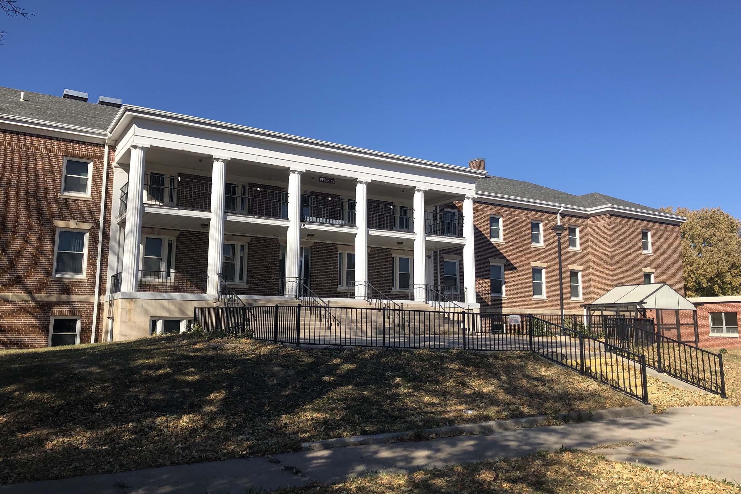 A brick building featuring a set of white columns at the entrance, with multiple balconies and large windows. There is a wide staircase leading to the front, surrounded by grass and scattered autumn leaves under a clear blue sky. The structure appears to have a historic or institutional design.