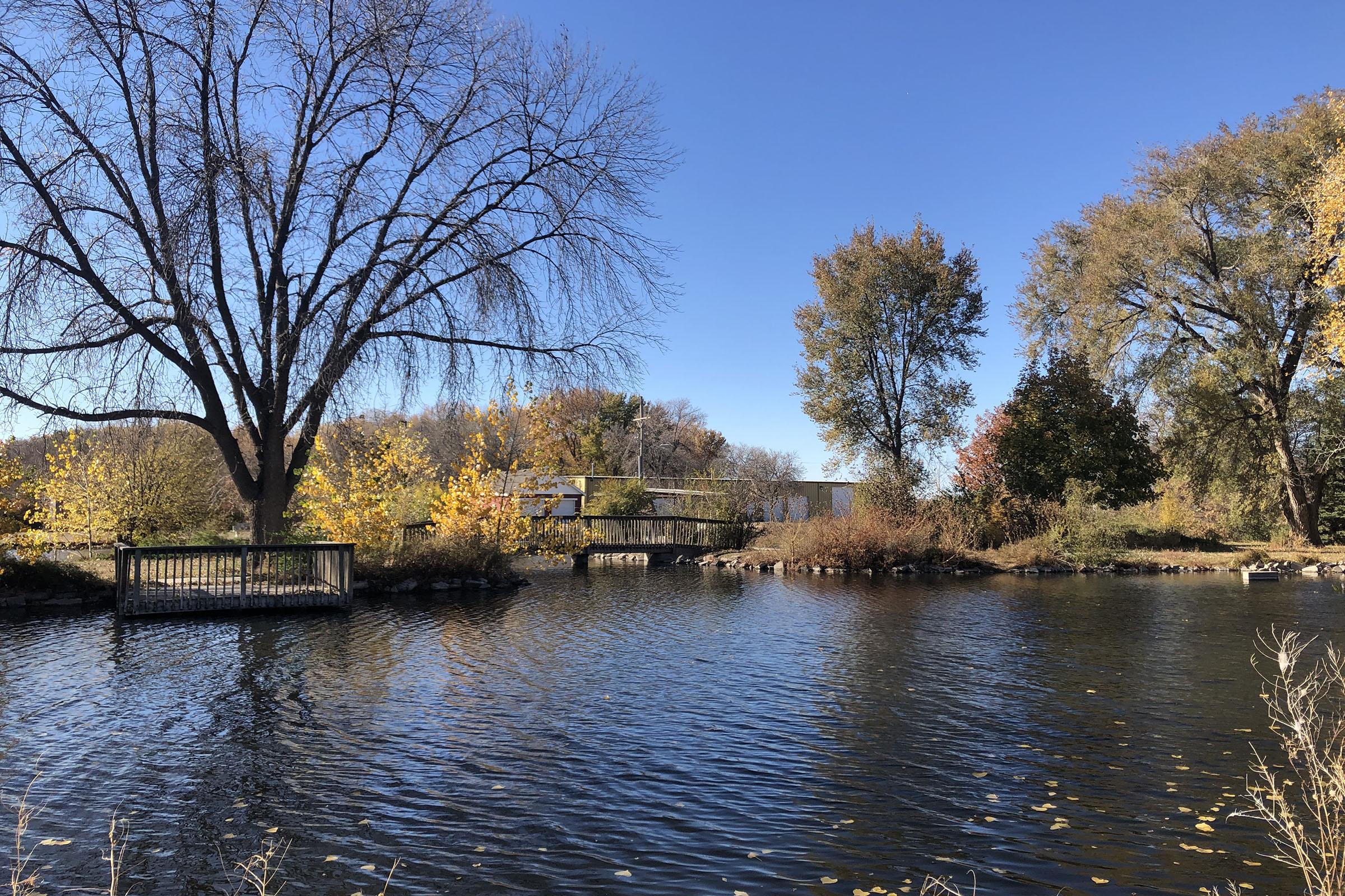 A tranquil scene featuring a calm river surrounded by trees displaying autumn foliage. The water reflects the clear blue sky, and a small wooden bridge can be seen crossing the river. The landscape showcases vibrant colors typical of fall, creating a serene natural environment.