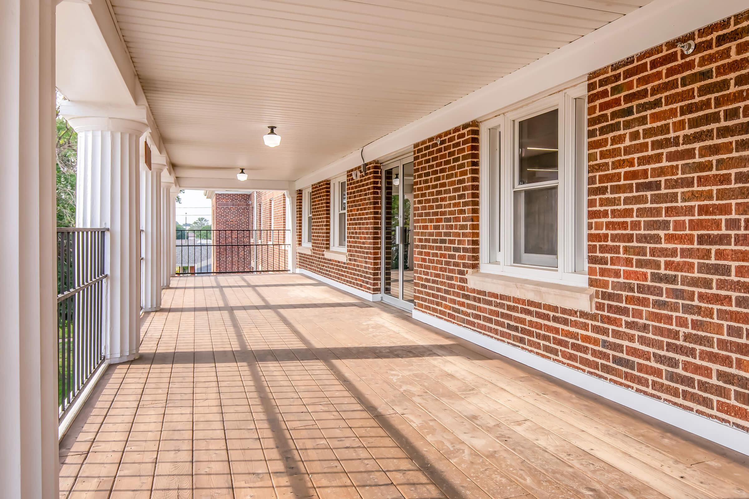 A spacious, covered balcony featuring a wooden floor, white columns, and brick walls. Natural light streams in through large windows. The area is clean and uncluttered, creating a welcoming outdoor space ideal for relaxation.