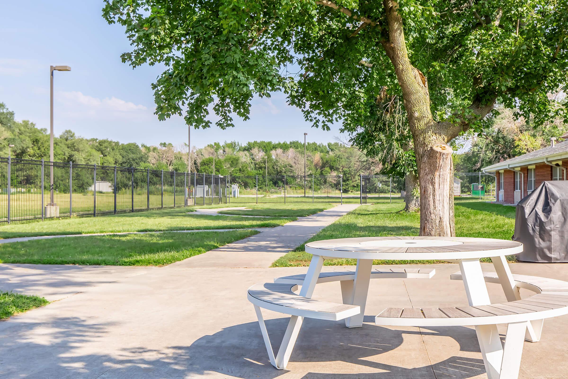 A white picnic table sits under a large tree in a park. In the background, a grassy area is enclosed by a black fence, with pathways leading further into the park. Several buildings can be seen on the right side, and the scene is bright and sunny, suggesting a pleasant outdoor setting.