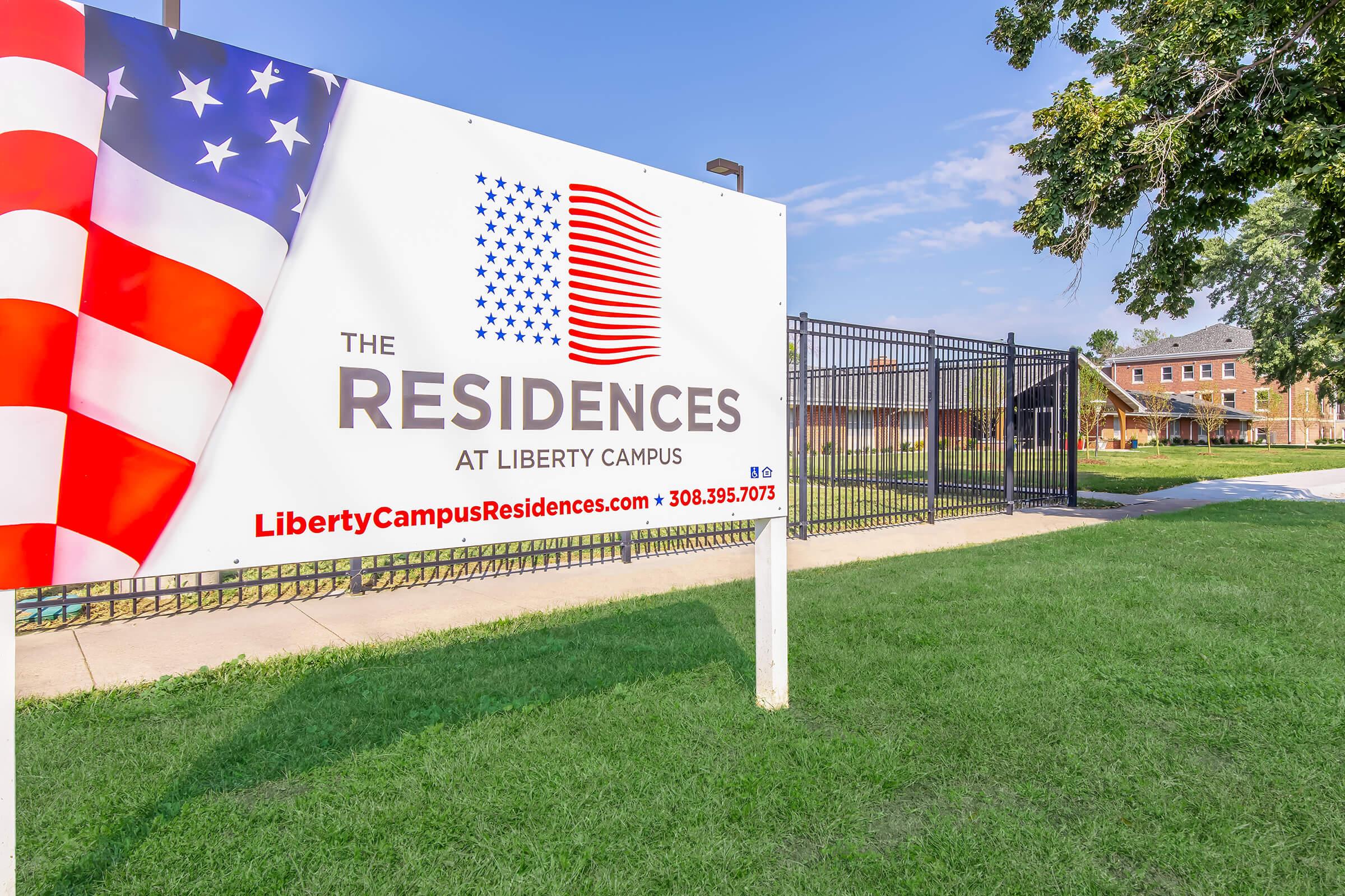 Sign for "The Residences at Liberty Campus," featuring an American flag design. The banner displays the website and phone number for inquiries, with a grassy area and fence in the background.