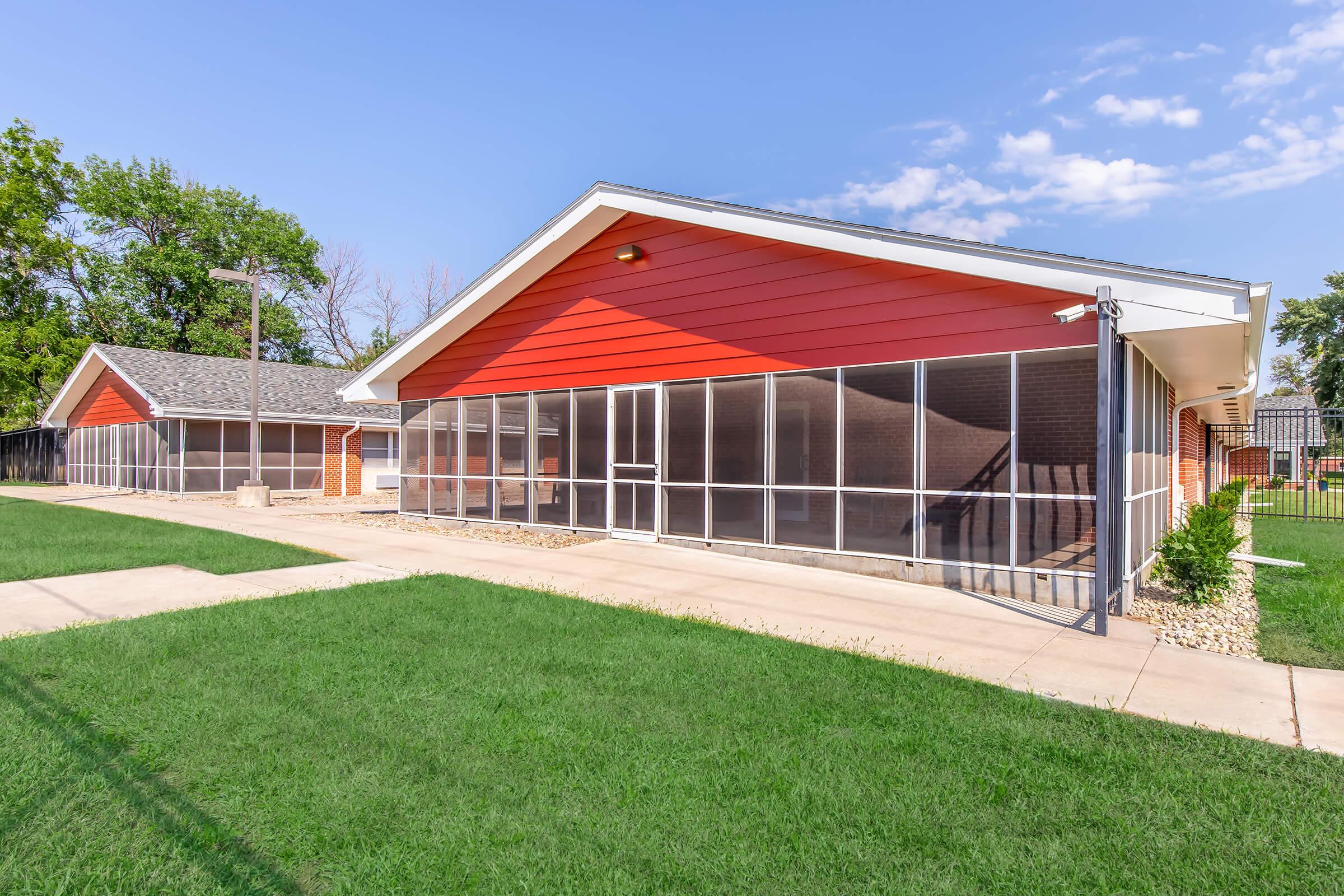 Modern single-story building with red siding and large screened windows, surrounded by well-maintained green grass. The building features a spacious outdoor area with walkways and a fence, under a clear blue sky with a few clouds.