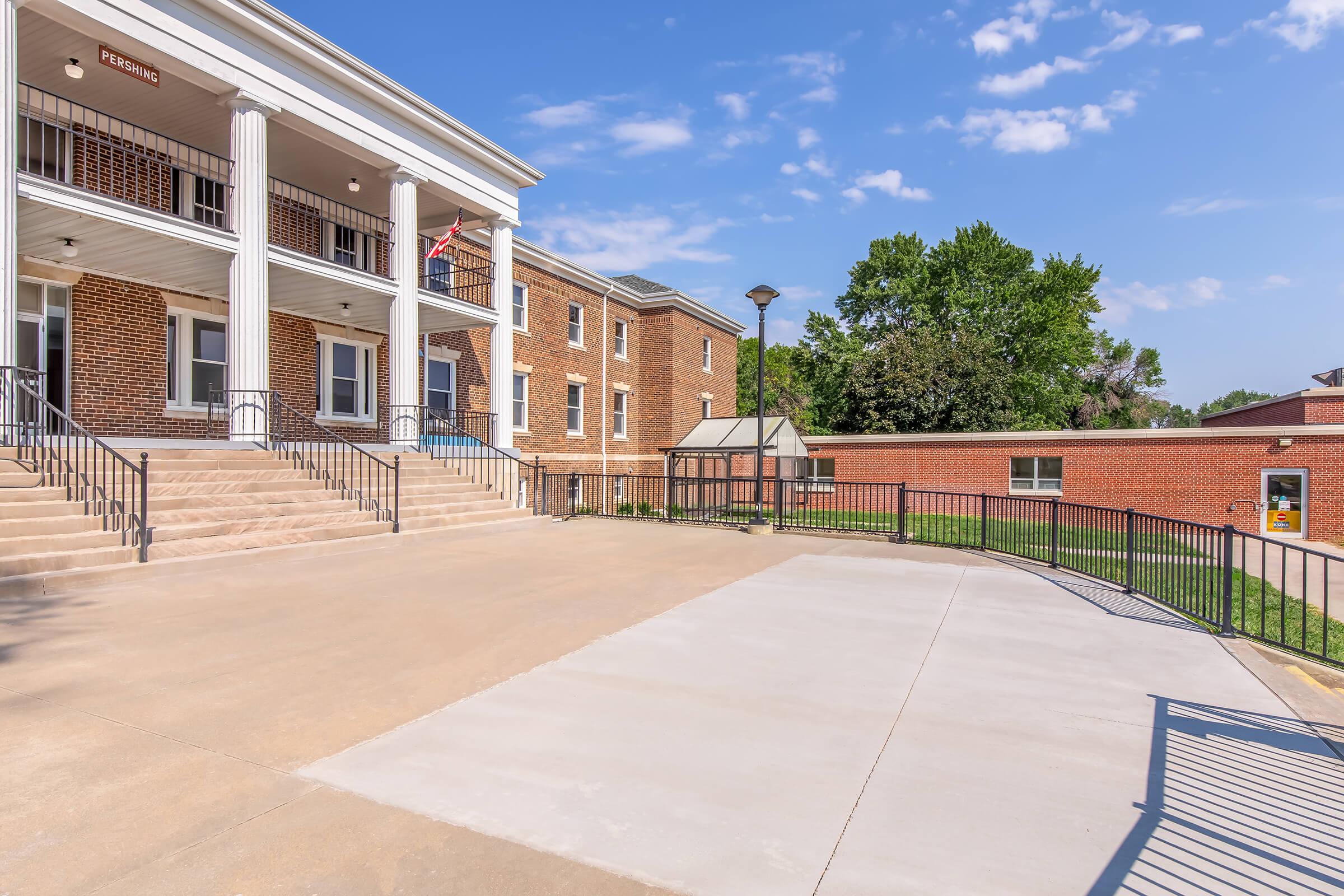 A view of a two-story brick building with white columns and a staircase leading up to the entrance. There are American flags displayed, and a well-maintained courtyard area in front. The sky is blue with scattered clouds, and a small, adjacent brick structure can be seen in the background.