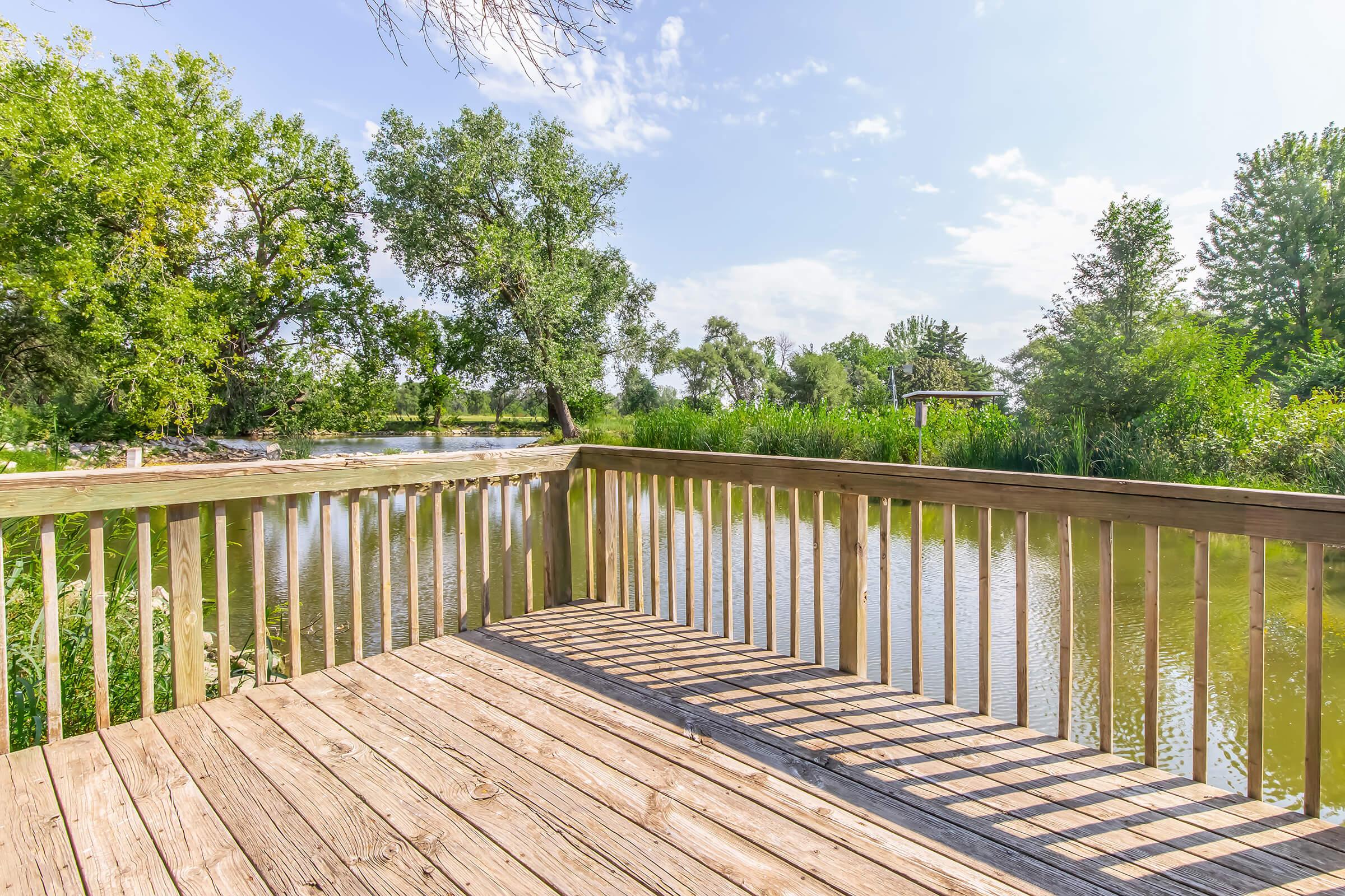 Wooden deck overlooking a calm river with lush green trees and plants on the banks. The scene is bright and sunny, with a few clouds in a blue sky. The deck has long shadows cast by the railing, creating a tranquil and inviting atmosphere. Perfect for relaxation and enjoying nature.