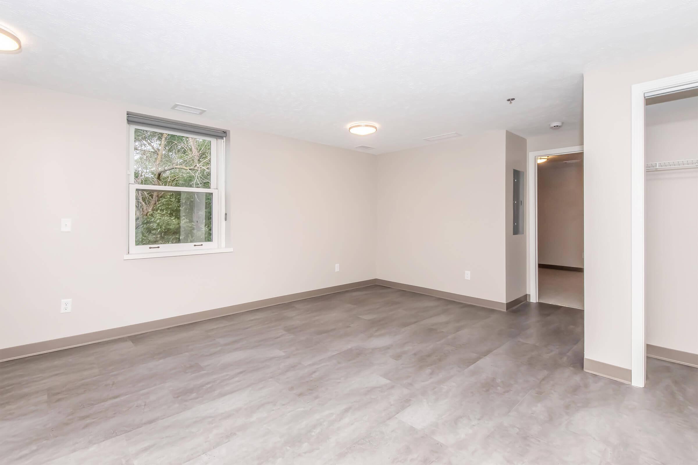 Empty room with neutral-colored walls and grey flooring. A window allows natural light, while a doorway leads to another area. The room has ceiling lights and no furniture, creating a spacious, minimalist atmosphere.