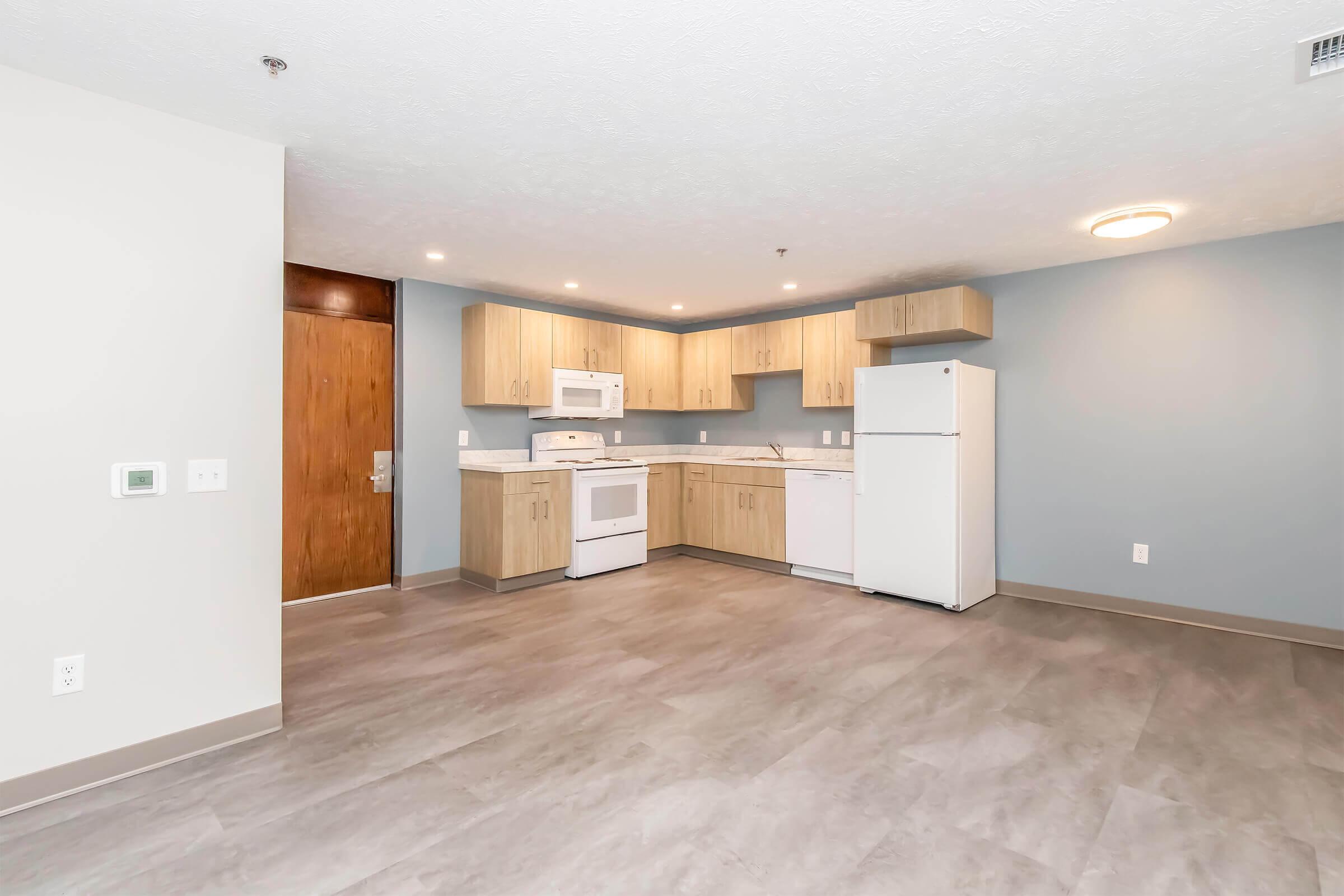 Spacious modern kitchen with light wood cabinetry, white appliances including an oven, microwave, and refrigerator. The room features a neutral-colored wall and a light gray floor, with ample natural light illuminating the space.