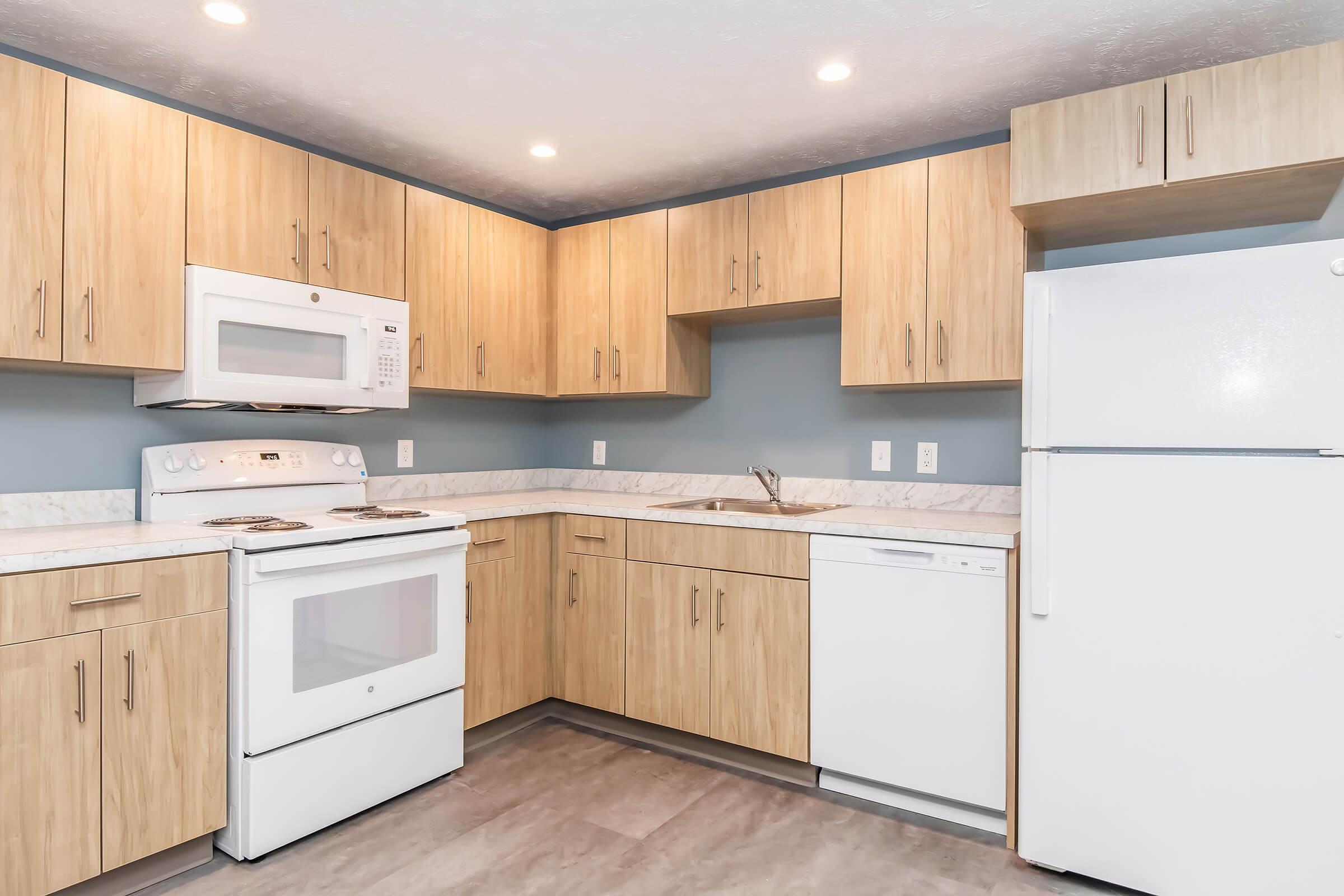 Modern kitchen featuring light wood cabinets, a white countertop, and stainless steel appliances. Includes a microwave, oven, and refrigerator, with a dishwasher beside the sink. Soft lighting enhances the inviting atmosphere. The walls are painted a subtle blue, complementing the cabinetry.