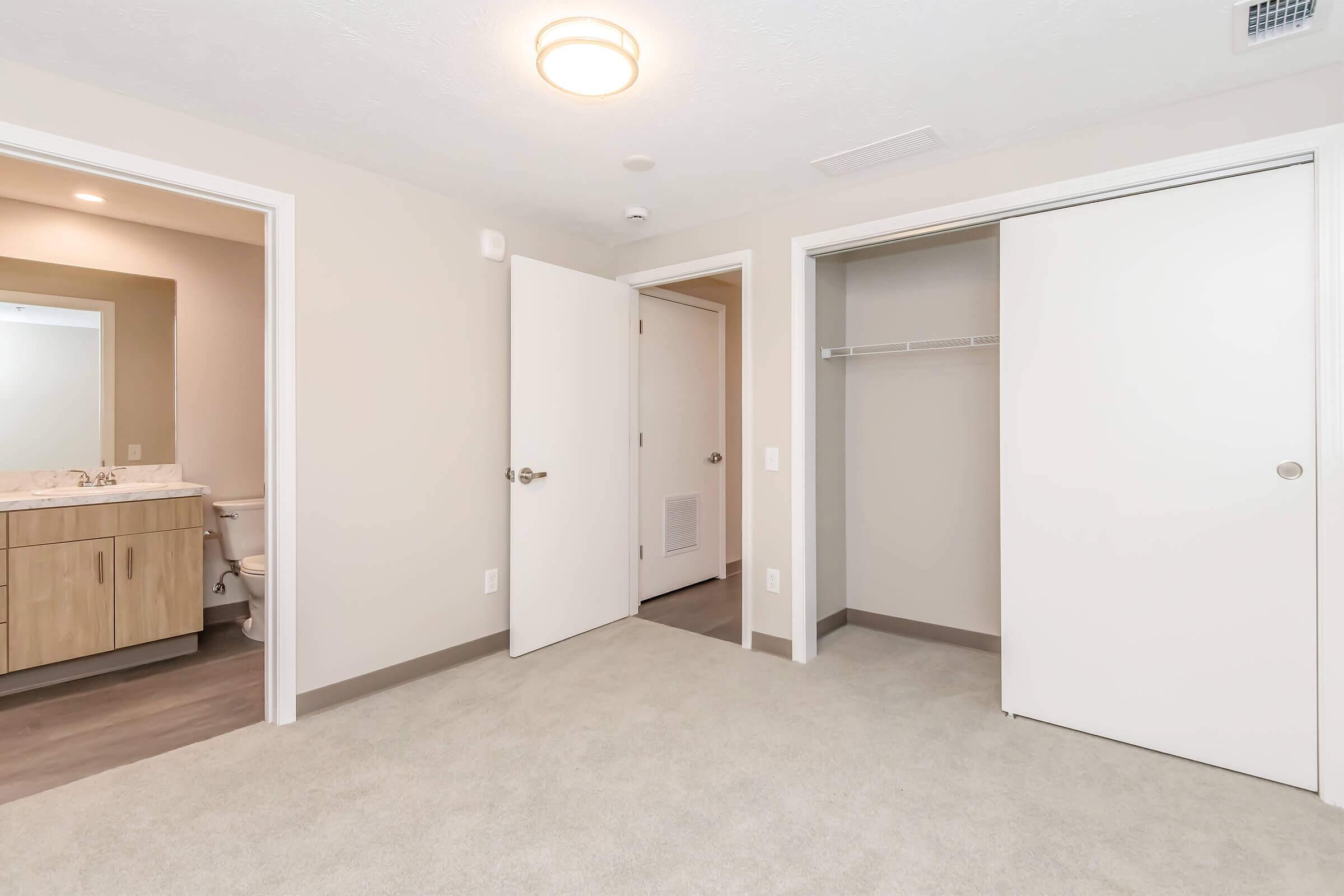 A spacious, empty room featuring light-colored walls and carpet. Two doors lead to a closet and a bathroom area, while a ceiling light provides illumination. The overall ambiance is clean and modern, suitable for a bedroom or living space.