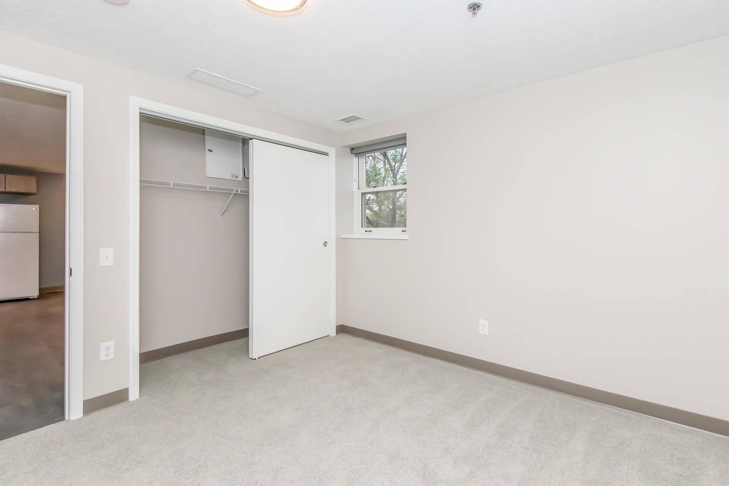 Interior view of a room featuring beige walls and carpet. A closed sliding door reveals a closet with a shelf. A window is visible, allowing natural light. The adjacent room is shown through an open doorway, with minimal furnishings and neutral decor typical of a rental space.