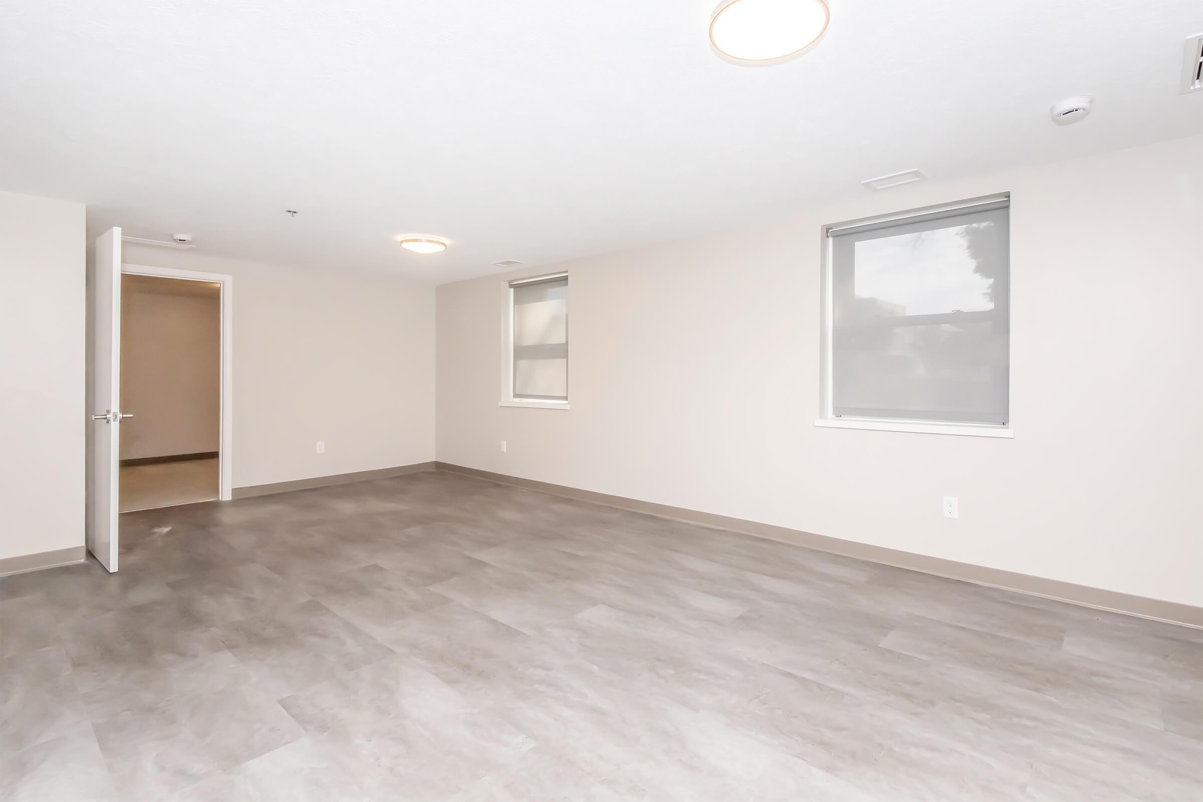 Empty room with light gray flooring, painted beige walls, and two windows providing natural light. A doorway leads to another room, and ceiling lights are visible. The space appears clean and modern, suitable for various uses such as living or office space.