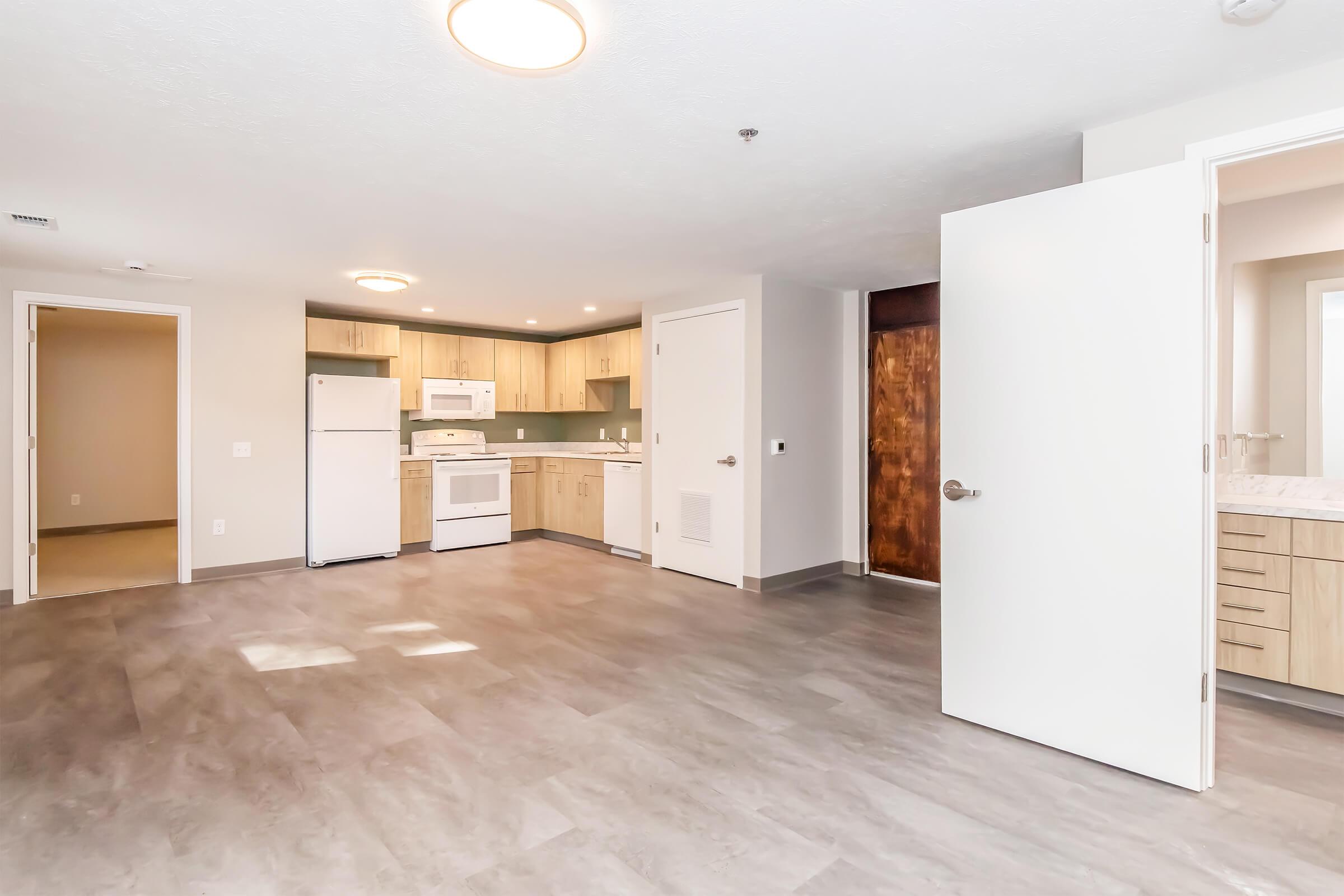 A bright and modern studio apartment featuring an open layout. The kitchen includes wood cabinetry, a white refrigerator, and stove. The living area has light-colored walls and flooring. A bathroom door is visible on the right, and there are two doors leading to other rooms. Soft lighting enhances the spacious feel.