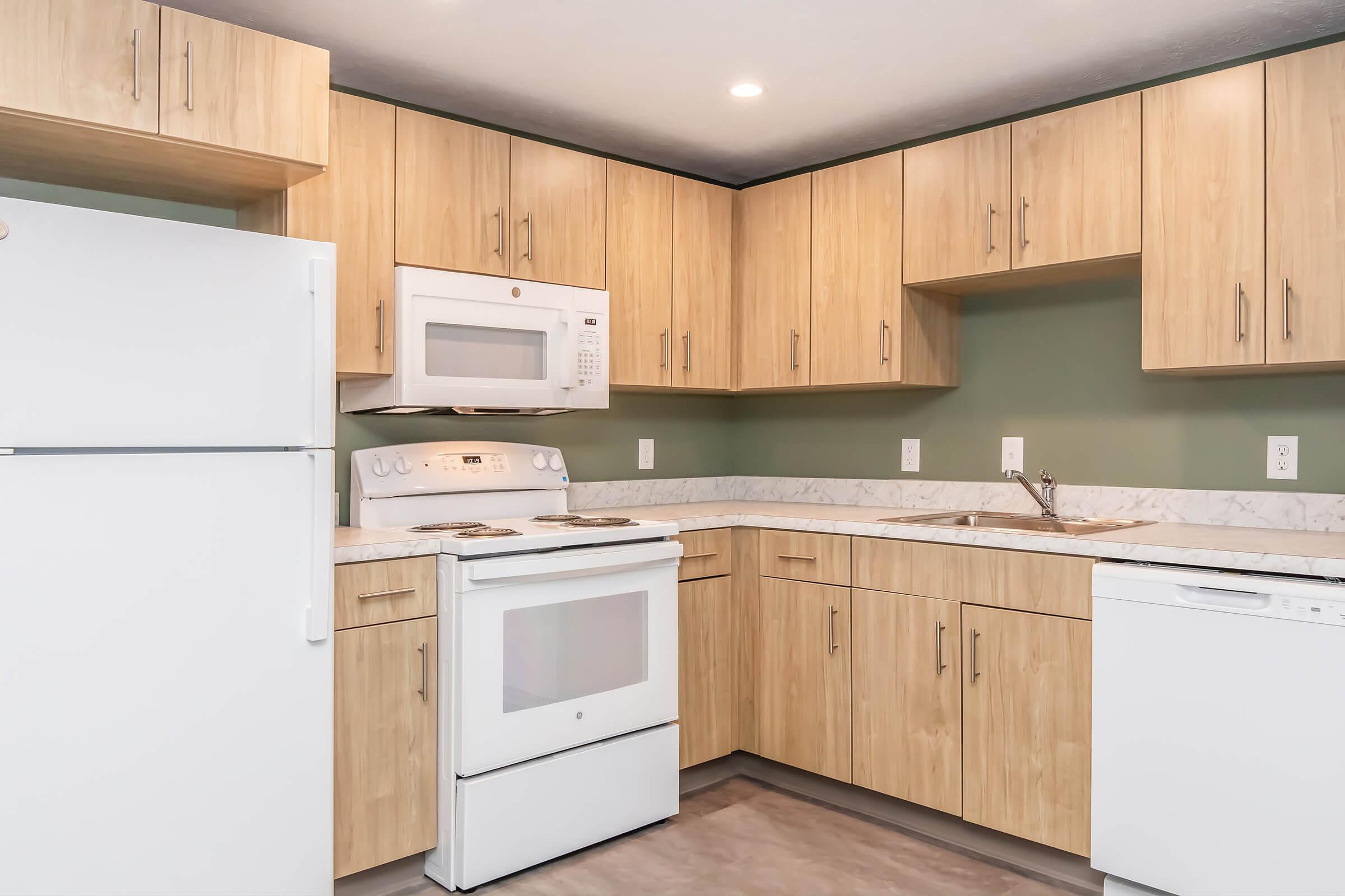 Modern kitchen featuring light wood cabinetry, a white refrigerator, a white stove with an oven, a microwave above the stove, and a white dishwasher. The countertops are a light marble pattern, and the walls are painted a muted green. The space is well-lit with recessed lighting.