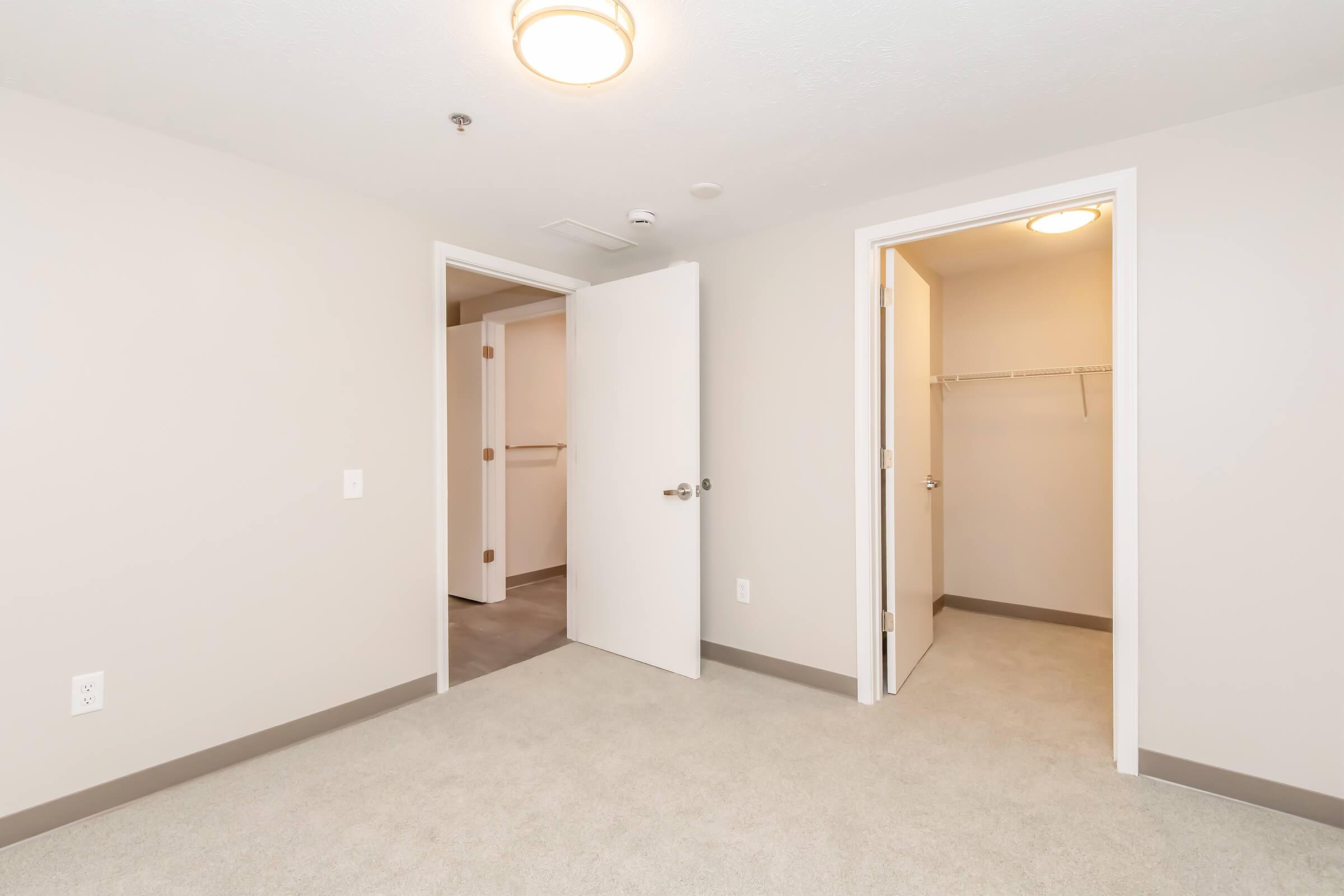 An empty room with light-colored walls and carpet. There are two doors, one leading to a closet and the other to another space. The ceiling has a round light fixture. The area is bright and uncluttered, showcasing a modern interior design.