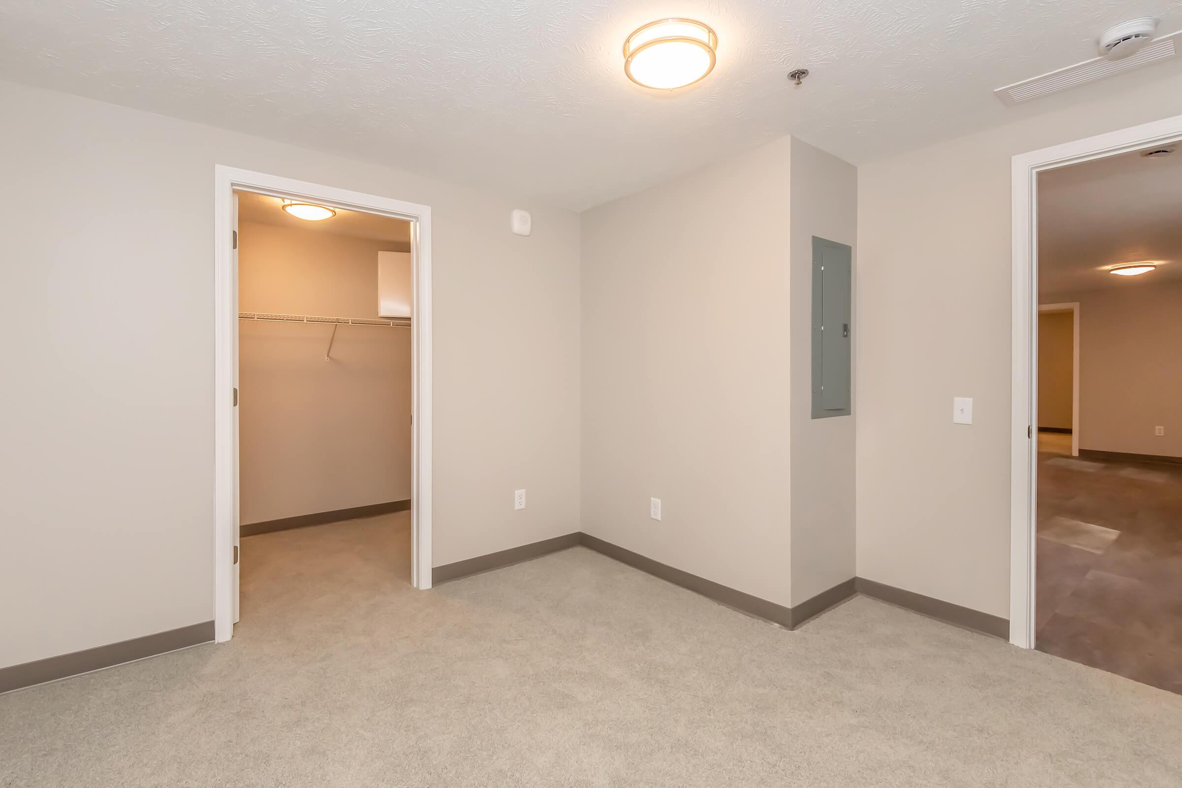 A light-filled room with beige walls and gray carpet. It features two doors, one leading to a closet with a hanging rod and the other opening to an adjacent room. A light fixture is mounted on the ceiling, and there is an electrical panel on the wall. The space is unoccupied and ready for furnishing.