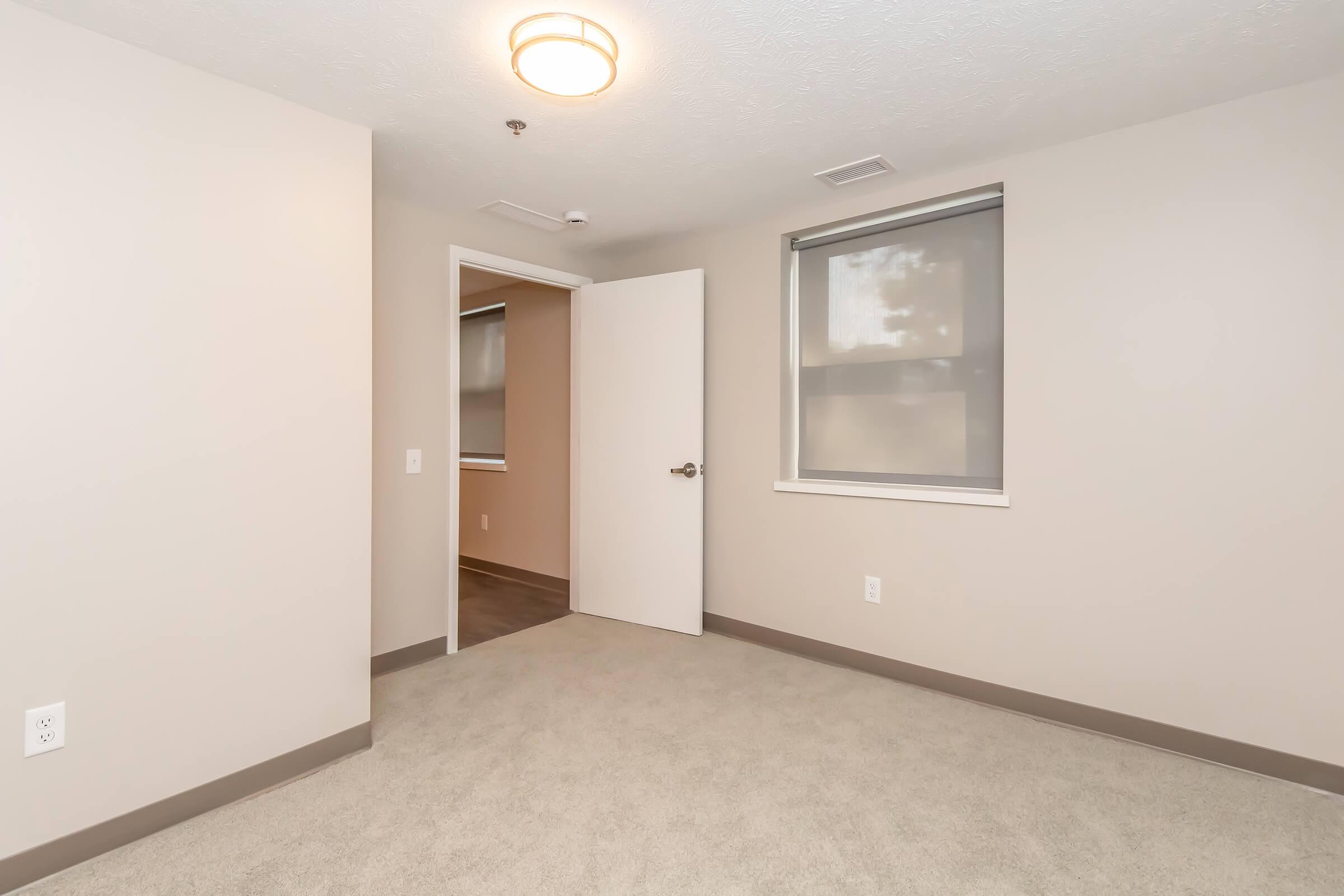 A well-lit, empty room featuring beige walls and a light-colored carpet. A simple ceiling light is installed, and there is an open door leading to another room. The windows are covered with light gray blinds, contributing to a clean and minimalistic aesthetic.