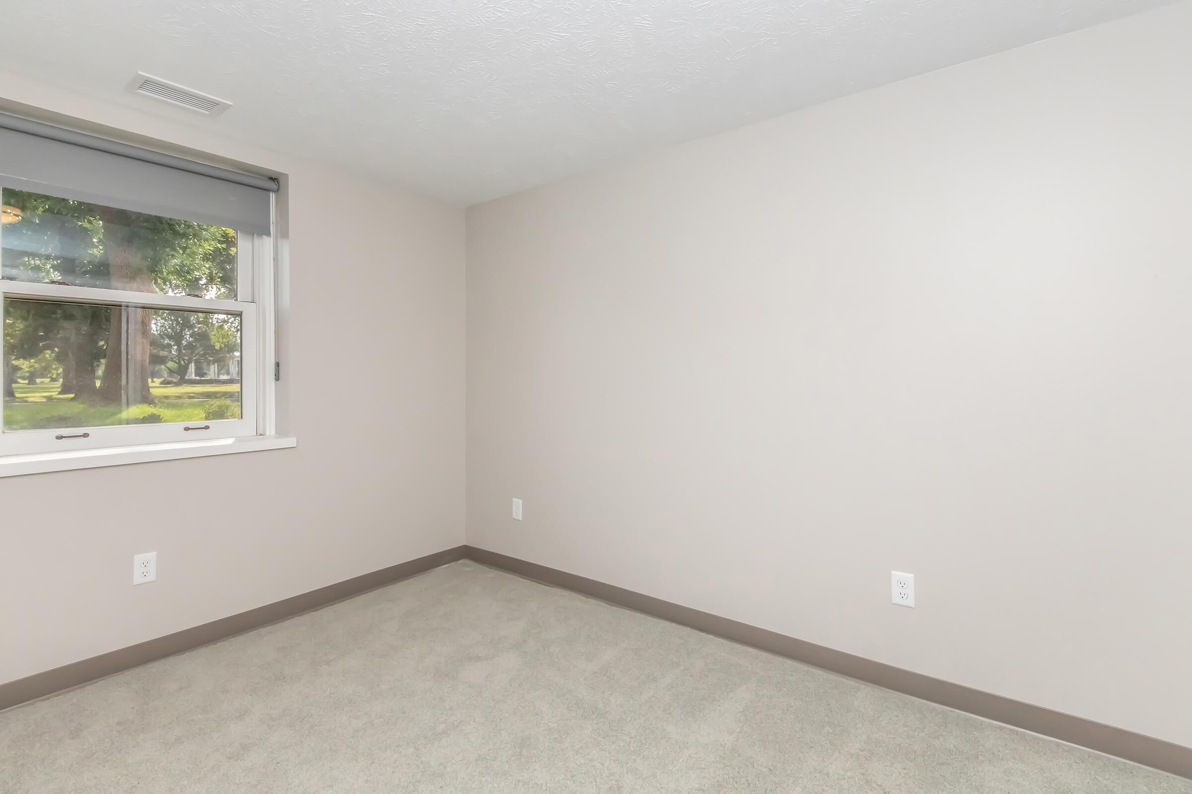 A light-colored empty room with beige walls and a soft carpet. There is a window on one wall, allowing natural light to enter. The view outside includes greenery. The room features minimal decor and is spacious, creating an open and airy feel.