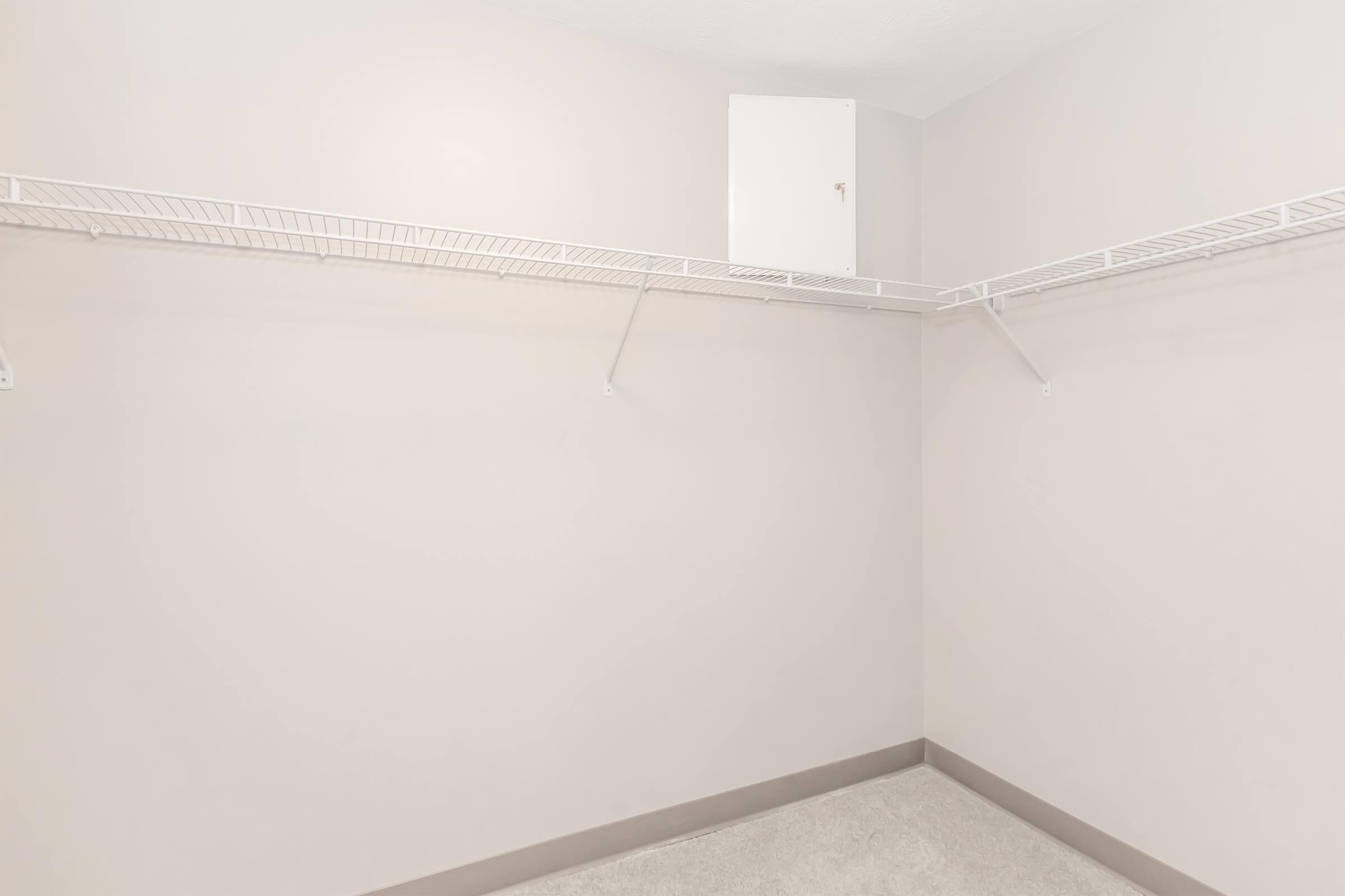 A spacious, empty closet with light beige walls and carpet. It features white wire shelving on two sides and a small hidden door near the ceiling, indicating additional storage space. The overall aesthetic is clean and minimalistic, emphasizing the room's potential for organization.