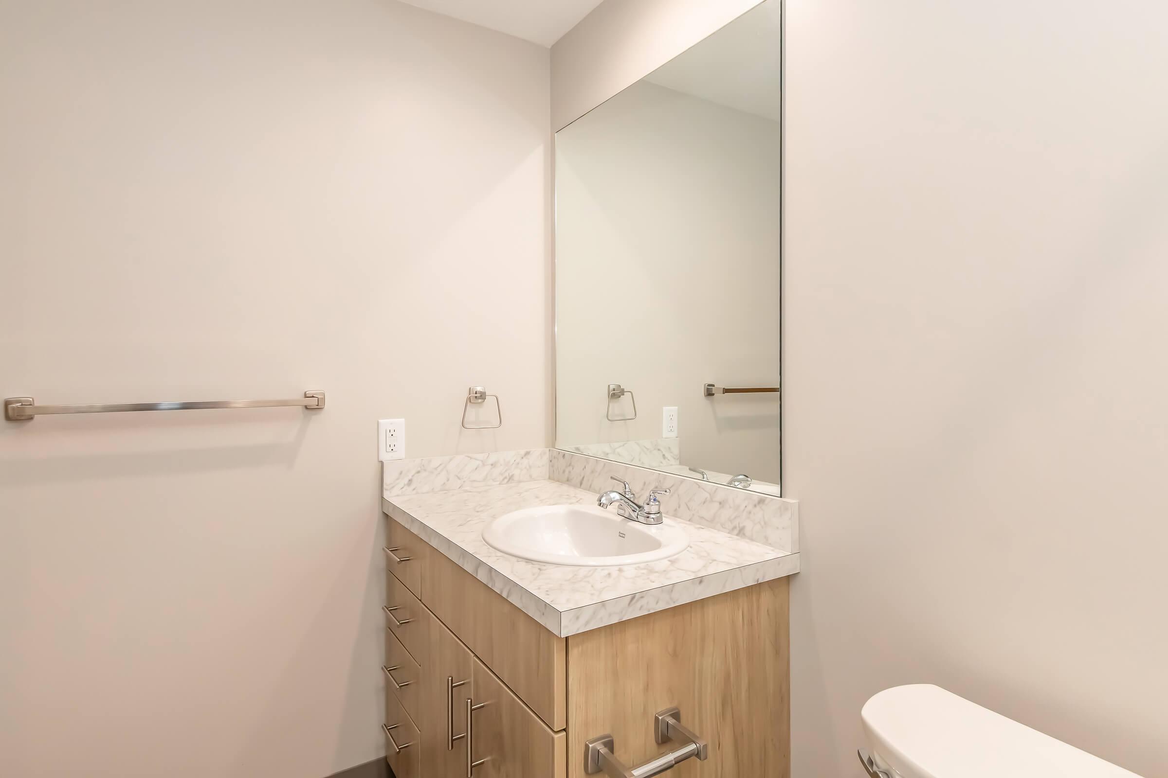 A modern bathroom featuring a light-colored wall, a large mirror above a white sink with a marble countertop, and wooden cabinetry. A towel bar is mounted beside the mirror, and a white toilet is visible to the right. The overall design has a clean and contemporary aesthetic.
