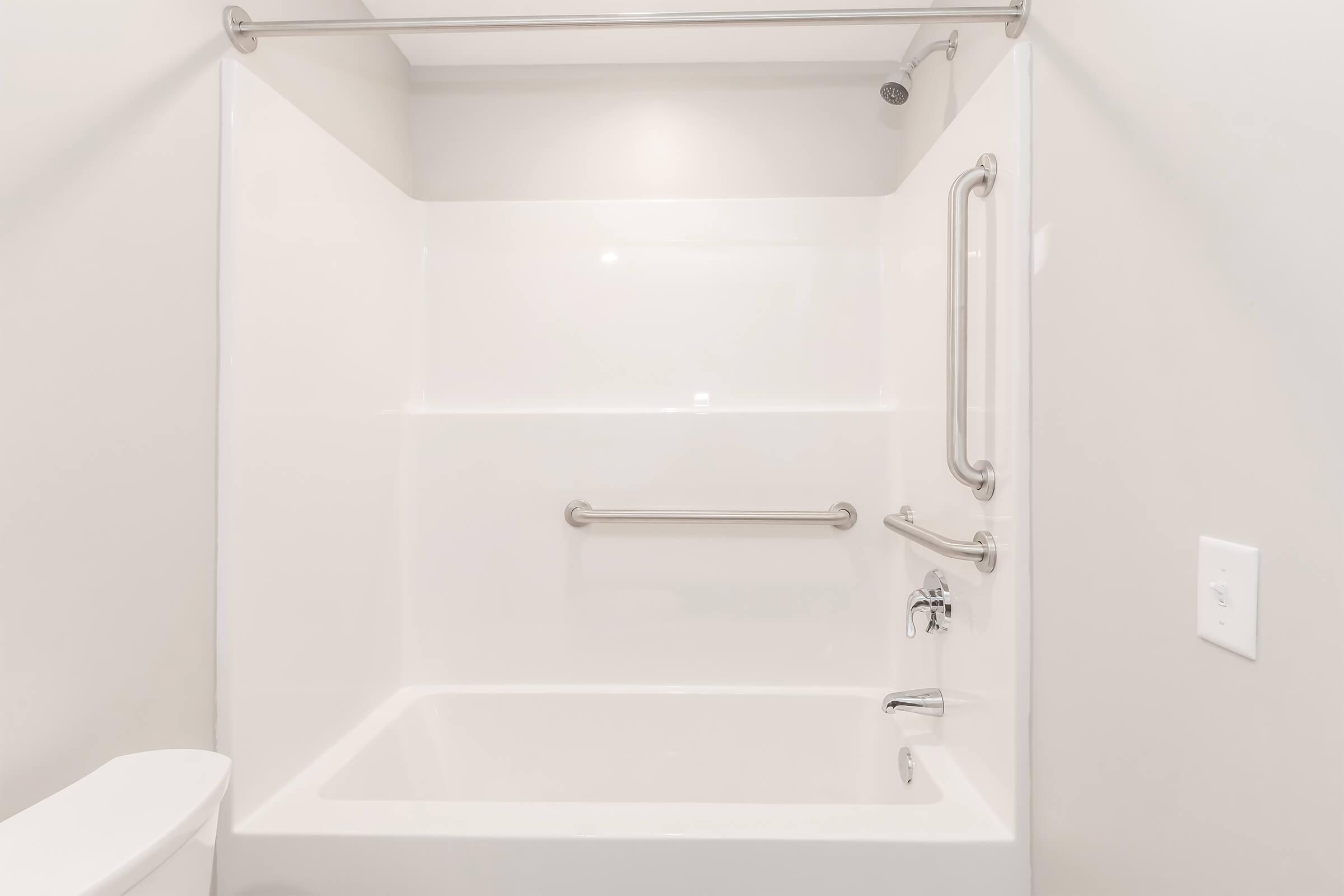 A clean, white bathroom with a bathtub and shower. The bathtub features a grab bar for safety, and there's a showerhead above. The walls are smooth and light-colored, with a simple toilet visible on the left side of the image.