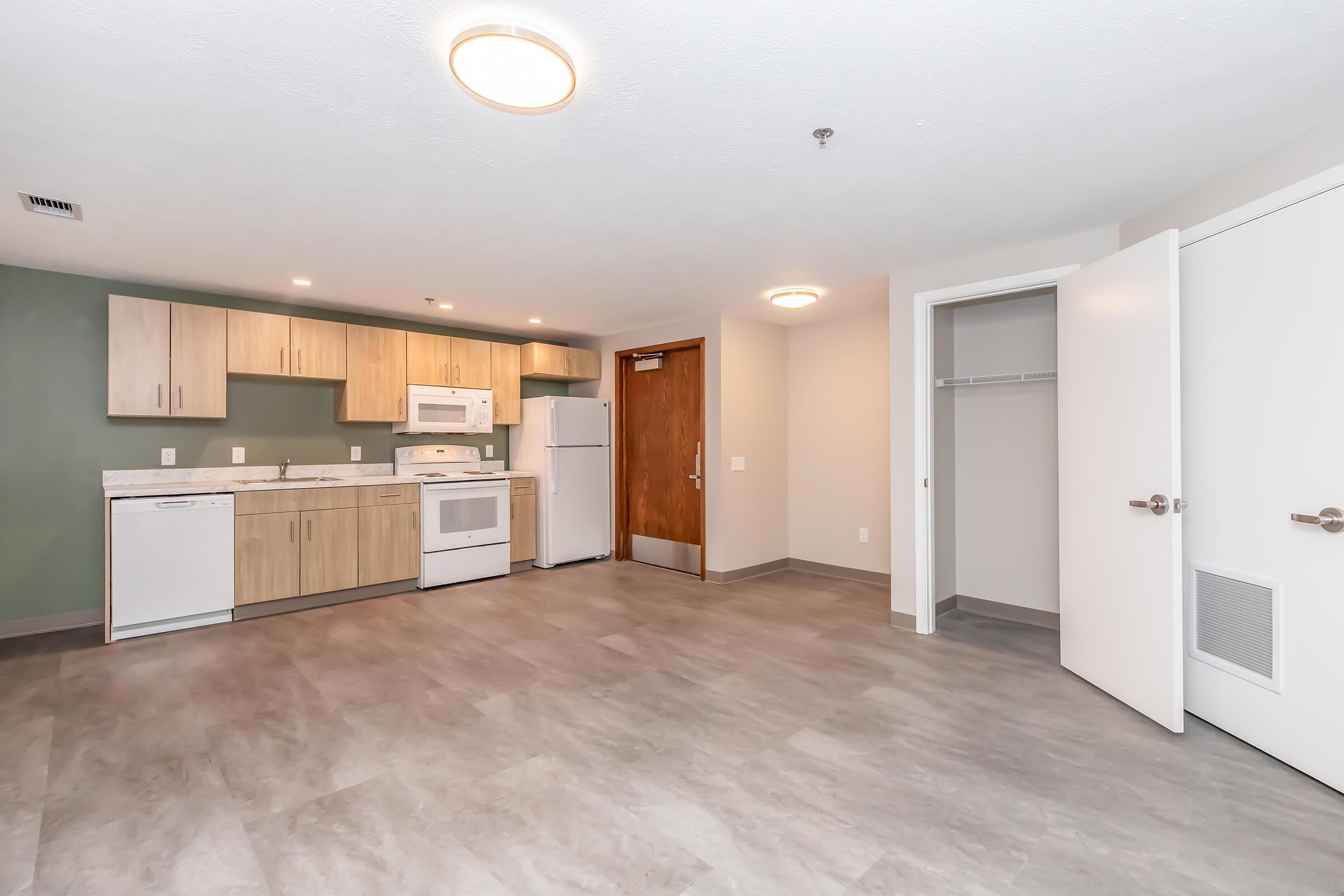 A modern, spacious apartment interior featuring a kitchen with light wood cabinets, a white microwave, stove, and dishwasher. The open layout includes gray flooring, a light fixture on the ceiling, a closet with a door, and a doorway leading to another room. The walls are painted in neutral colors.
