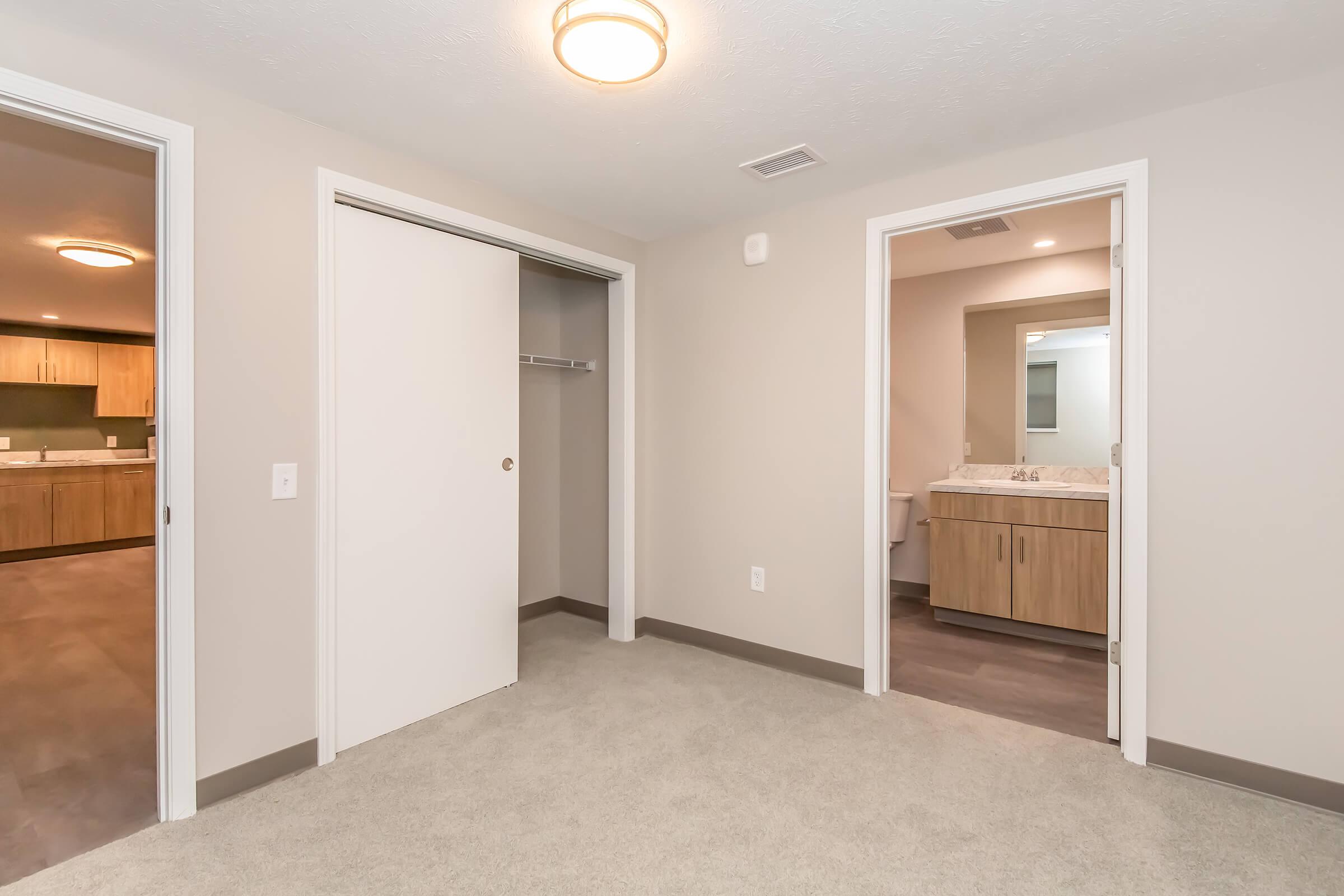 A spacious, neutral-colored room with carpeted flooring. To the left, there is an open door leading to a closet. On the right, a door opens to a bathroom with a wooden vanity and a mirror. Soft, overhead lighting illuminates the space, creating a warm atmosphere.