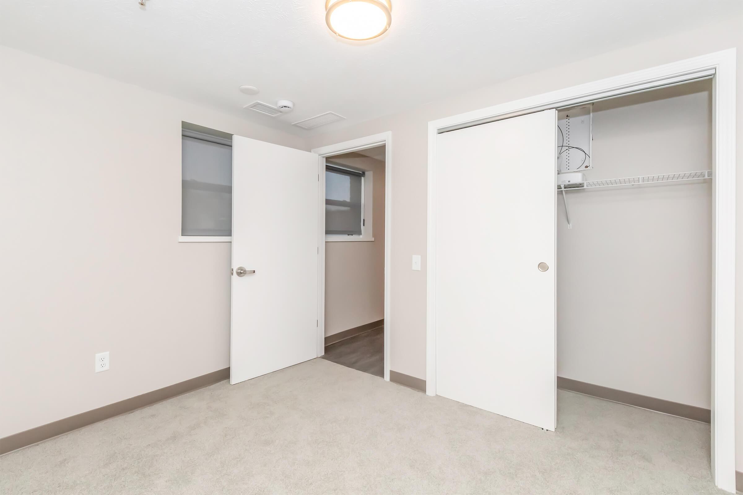 A beige room with light gray carpet, featuring two white doors: one leading to a closet with a shelf and the other leading to another room. Two windows are visible, with natural light coming in. The walls are painted a light color, creating a bright and airy atmosphere.