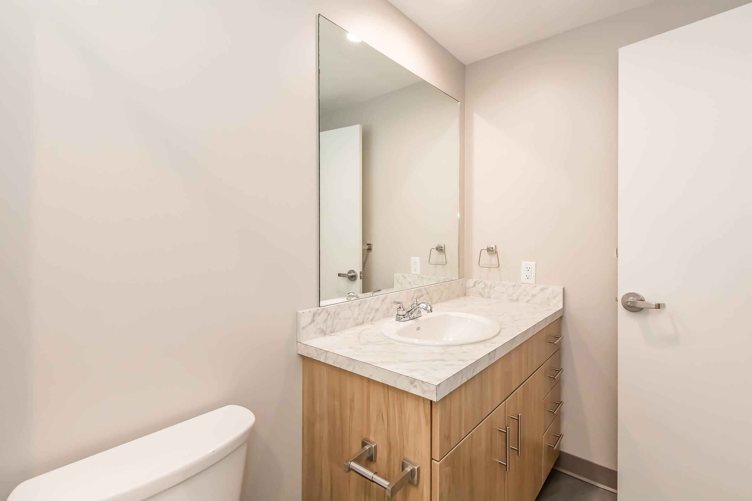 Modern bathroom with a light-colored wall, a large mirror above a white sink set in a wooden vanity with multiple drawers, a silver faucet, and a white toilet. The space is well-lit, featuring simple, clean lines and a minimalist design. A closed door is visible on the right.