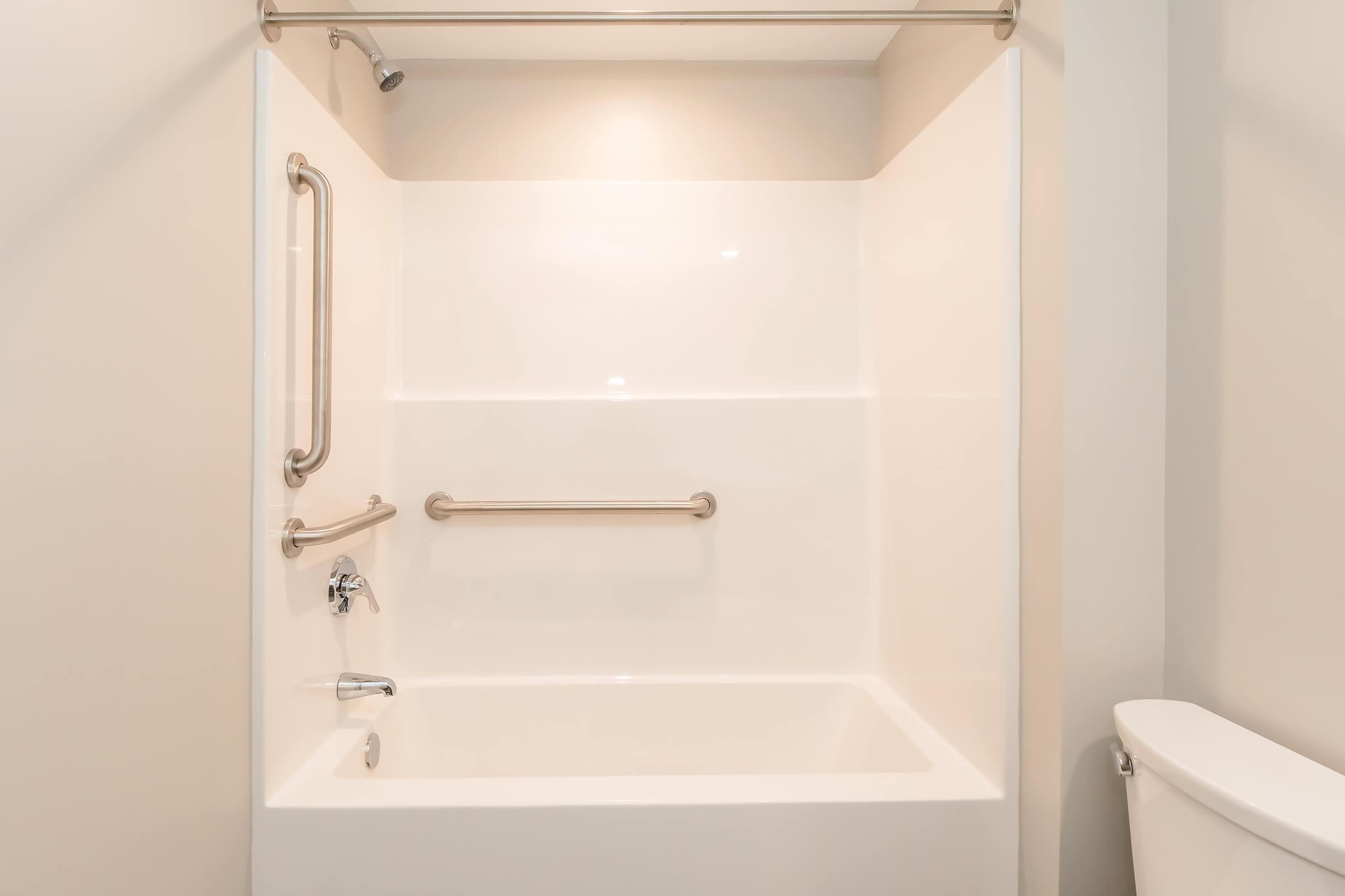 A clean, modern bathroom featuring a white, smooth-walled shower and bathtub combination with grab bars for safety. The shower has a chrome faucet, and a white toilet is visible in the corner. The space is well-lit with soft overhead lighting.