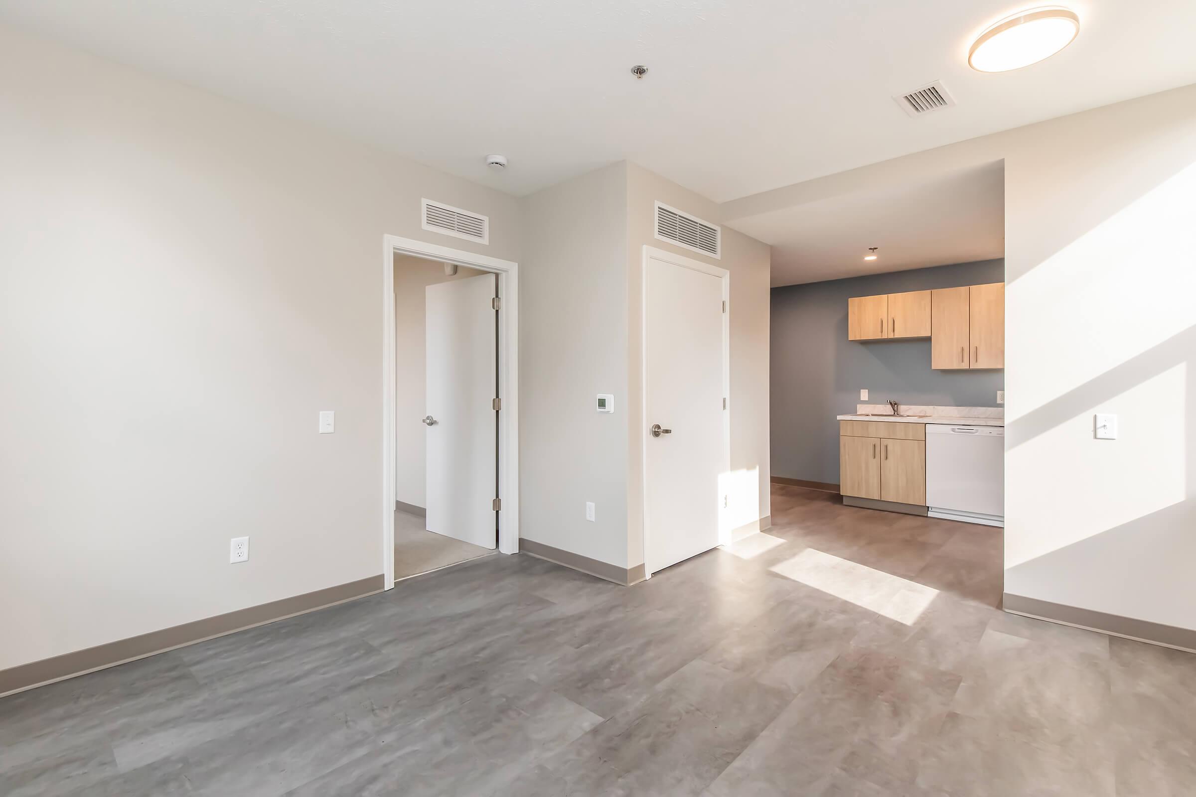 A modern, empty apartment living space featuring beige walls and gray flooring. On the left, a door leads to another room, and on the right, an open doorway leads to a kitchen area with light wood cabinetry and a white refrigerator. Natural light streams in, creating a bright atmosphere.