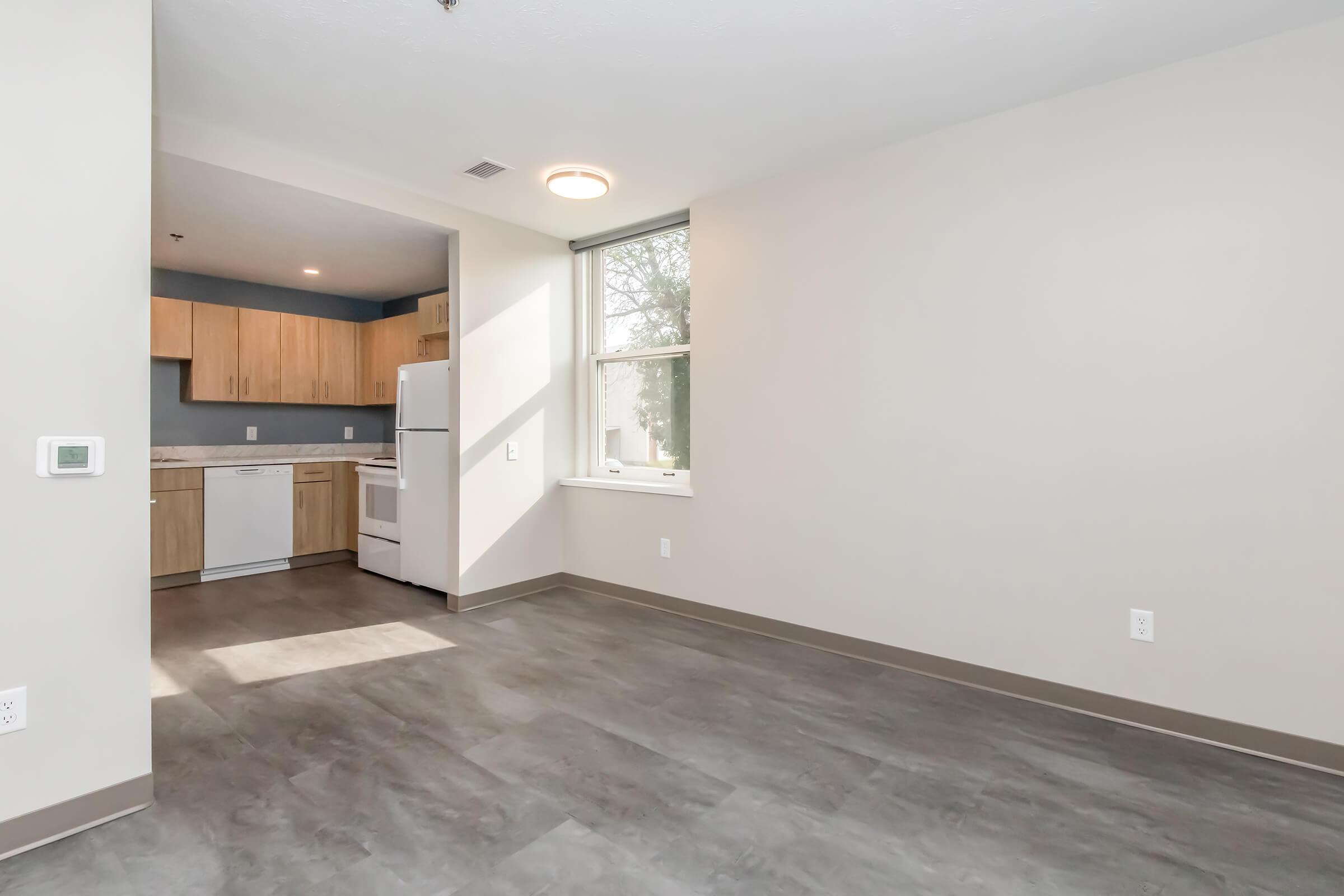 Interior view of a modern, unfurnished apartment featuring large windows, light-colored walls, and a gray floor. In the background, a compact kitchen with wooden cabinets, a refrigerator, and a stove is visible. The space is bright and airy, with natural light flooding in from the windows.
