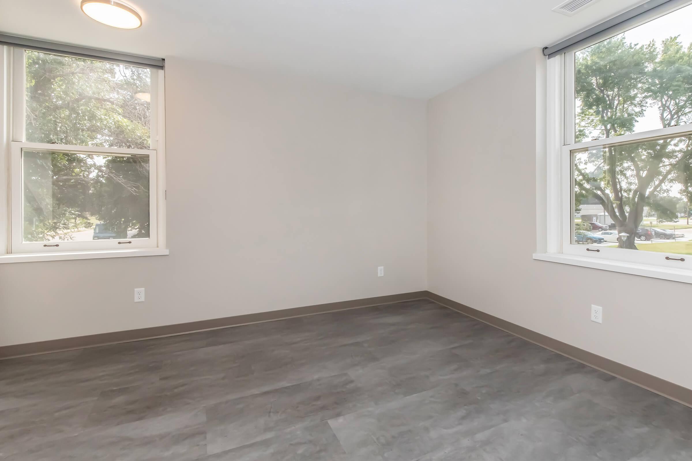 Empty room with gray flooring, pale walls, and two large windows providing natural light. One window offers a view of trees outside. The ceiling features a light fixture. The overall space is clean and minimalist, suitable for various uses.